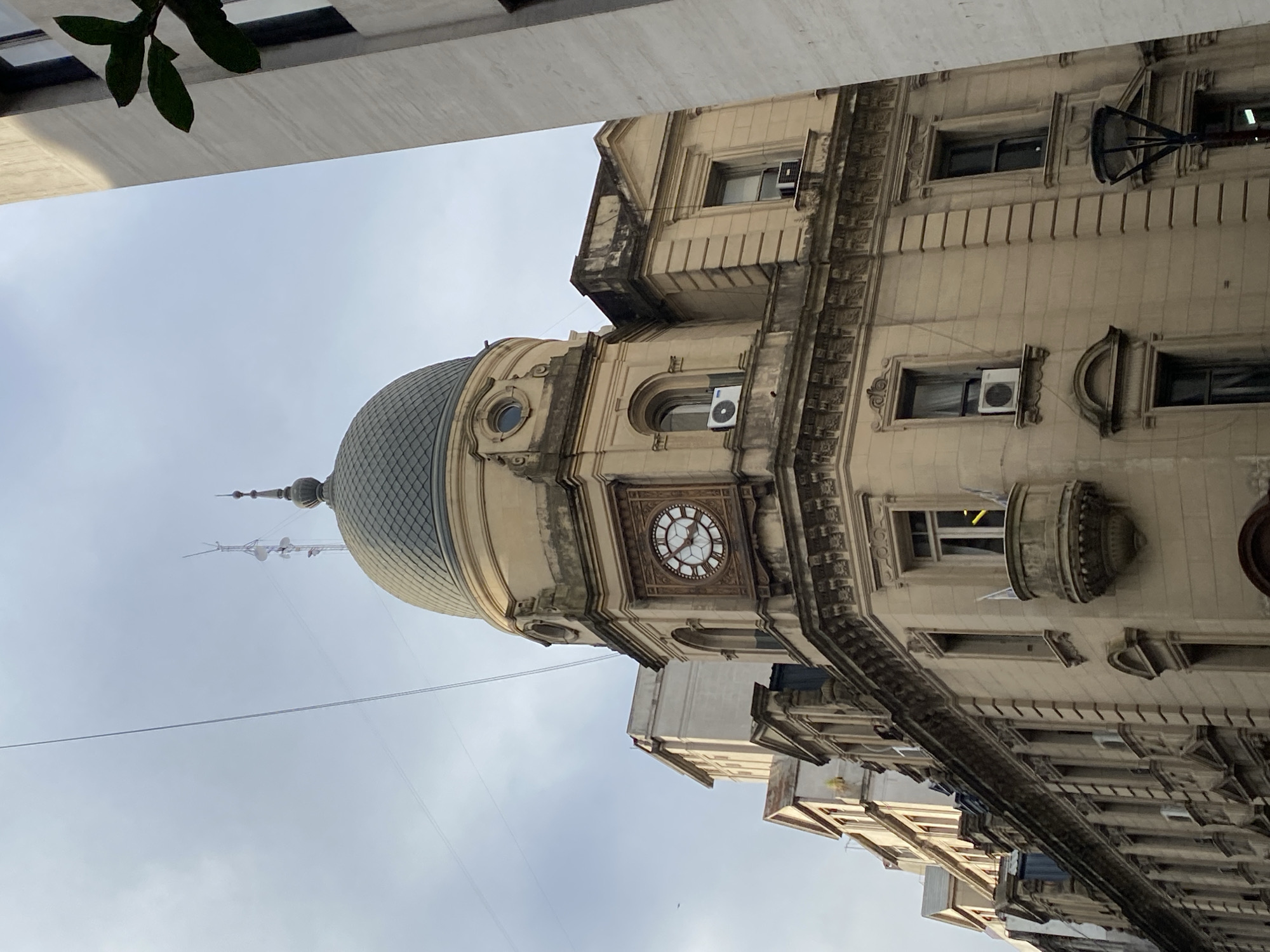 The image depicts a low-angle view of a Beaux-Arts style building with a prominent dome, likely located in Buenos Aires, Argentina. The building features a clock tower element and detailed architectural ornamentation, including moldings, window frames, and decorative stonework. Several windows have air conditioning units installed. The sky is overcast. Foliage is visible in the upper left corner of the image. Other buildings can be seen extending down the street.