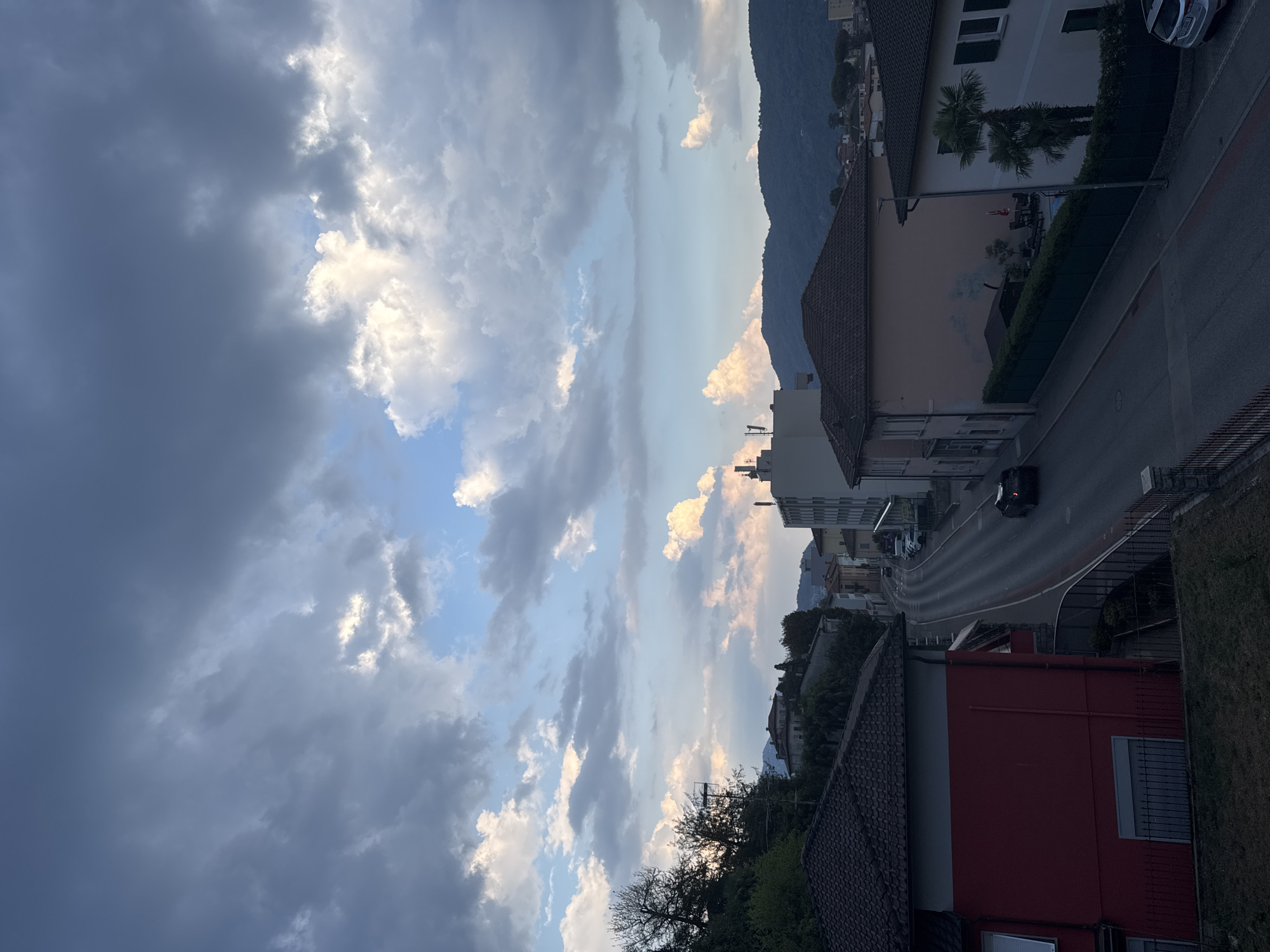 The image shows a view of a street in Balerna, Switzerland, from an elevated perspective. The sky is filled with dramatic clouds, some dark and grey, others illuminated by sunlight, suggesting an overcast or transitioning weather condition. The street is paved and curves through a residential area. On either side of the street are houses with tiled roofs, typical of European architecture. Some houses are painted in vibrant colors like red and pink, while others are lighter in shade. Greenery, including trees and bushes, is visible, especially on the left side of the image and in front of some houses. A few cars are parked or driving on the street. In the background, rolling hills or mountains are visible, adding to the scenic landscape. The overall impression is of a quiet, European town on a day with dynamic weather.