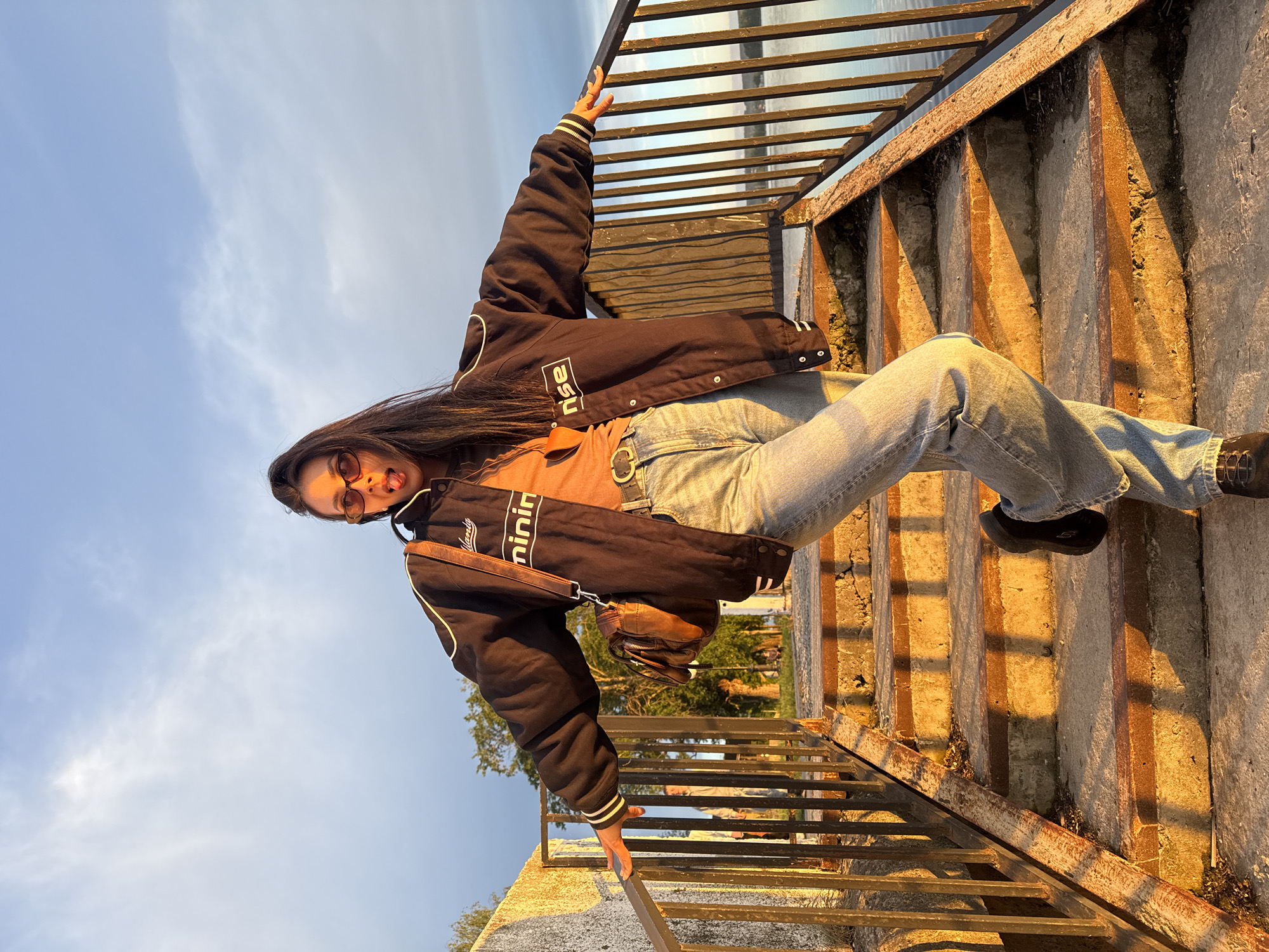 A young woman poses for a photo on an outdoor staircase in Votkinsk, Russia, with a body of water visible in the background. She is wearing a black bomber jacket, a brown t-shirt, and blue jeans. She is wearing sunglasses and sticking out her tongue. She is holding onto the railing with one hand and is seated on the steps. The staircase is made of concrete and metal. The lighting suggests it is near sunset. A brown purse hangs from her shoulder.