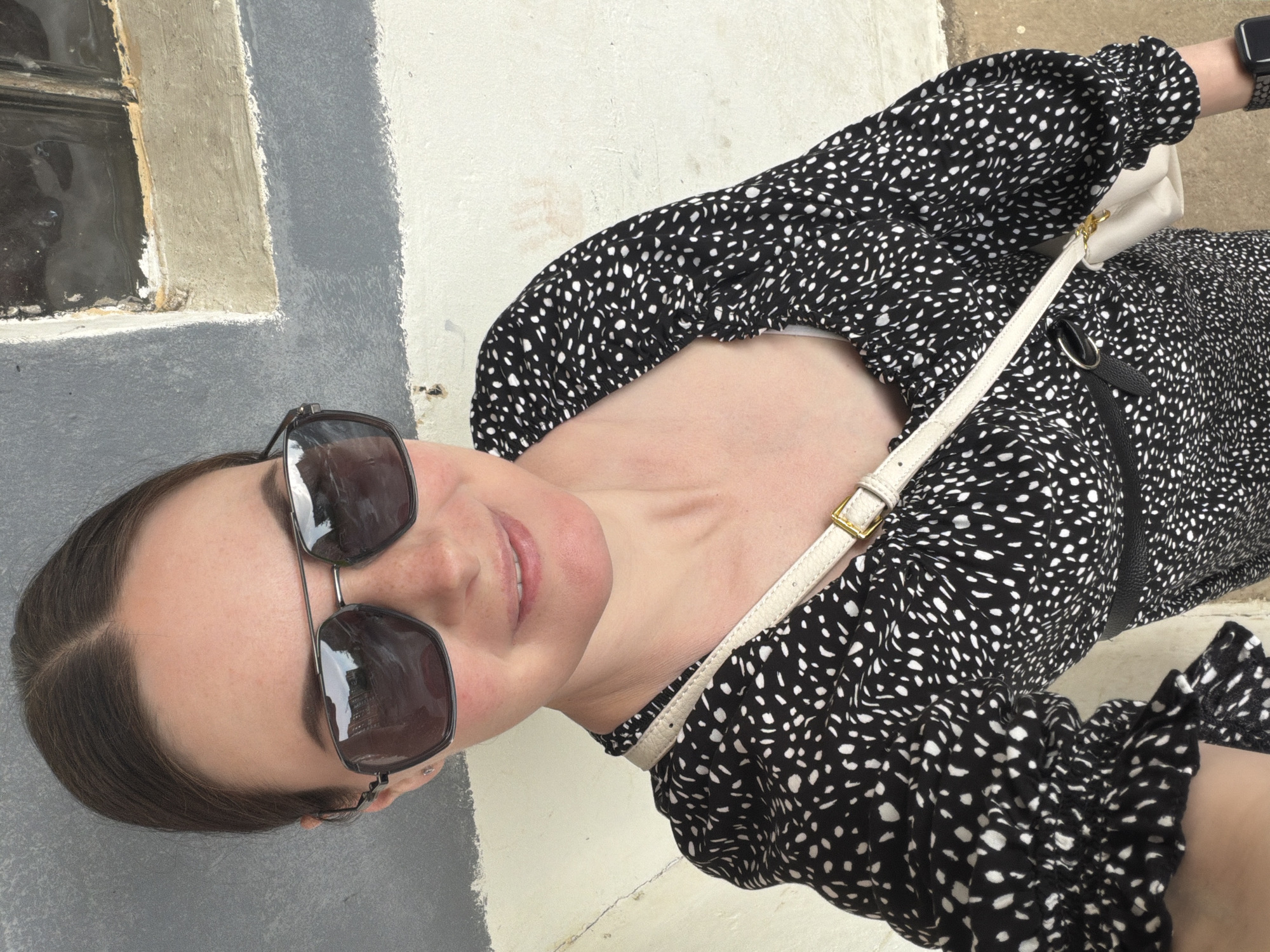 A woman with brown hair and sunglasses is wearing a black and white patterned dress with a white shoulder bag. She is standing near a building with a gray and white exterior. The location is in the city of Unknown, Unknown.