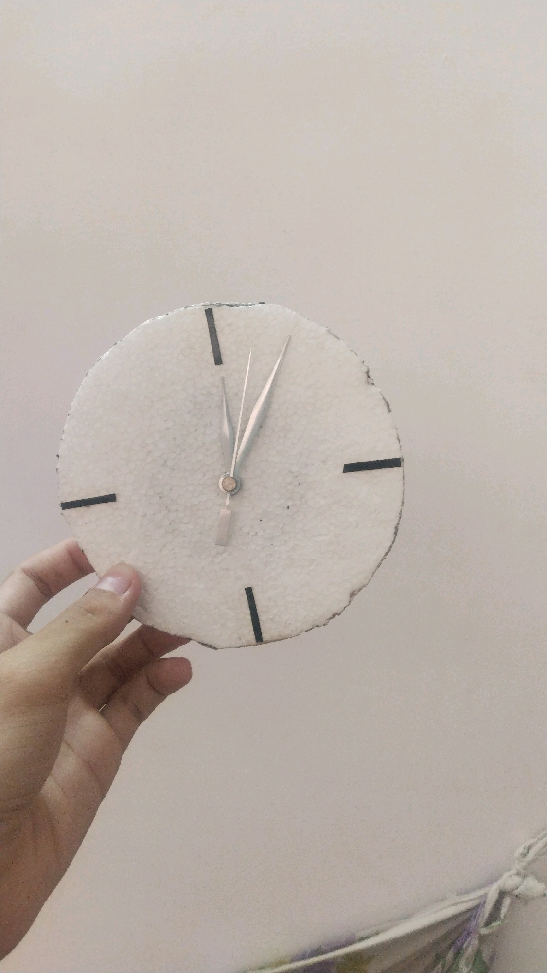 A close-up, slightly blurry shot shows a hand holding a handmade clock. The clock face is made of a circular piece of white material, possibly styrofoam, with black marks indicating hours. Two metallic clock hands are visible, pointing to approximately 11:05. The background is a plain, light-colored wall. The image is taken indoors. The location is not discernible from the image itself.