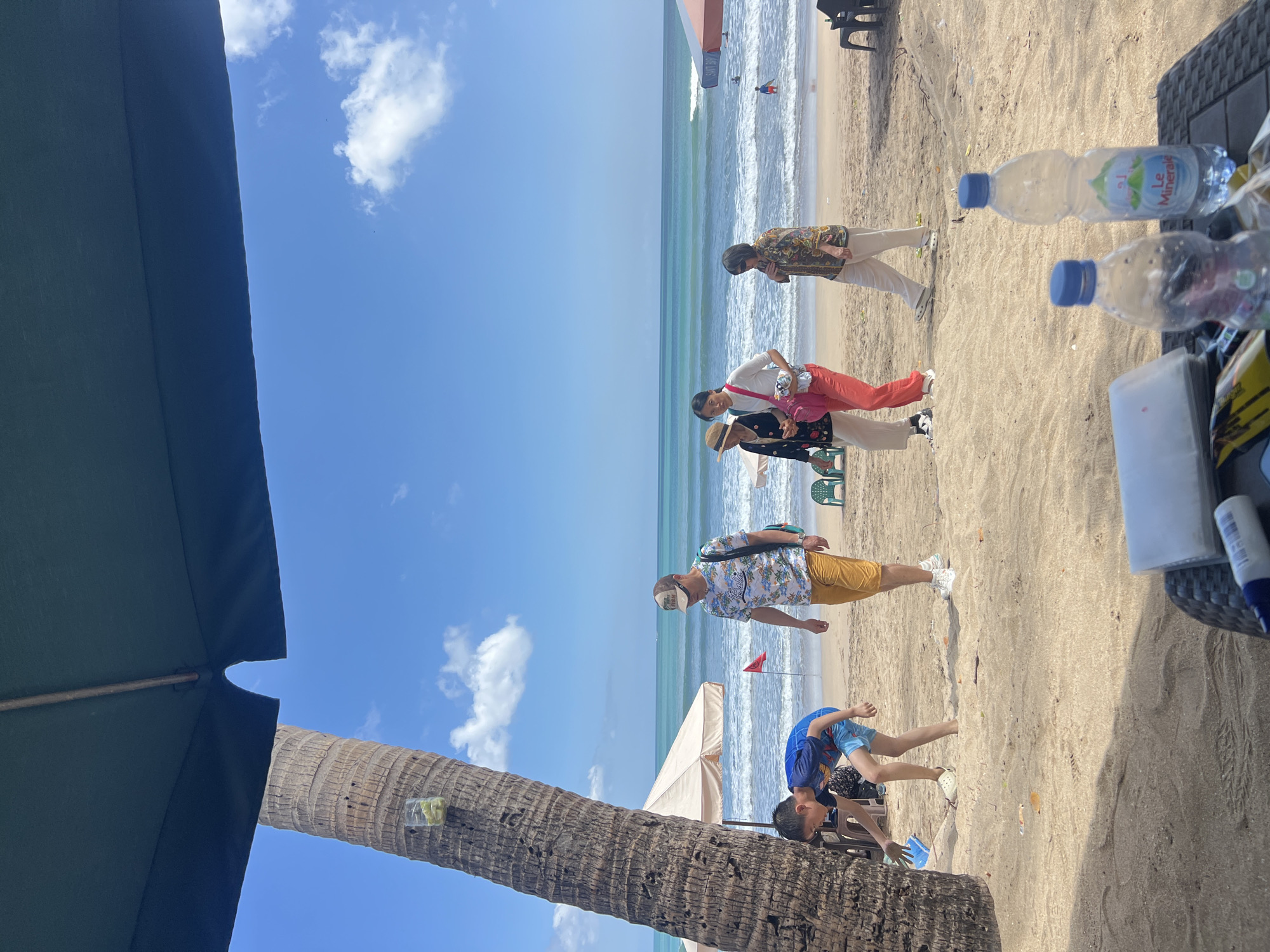 In Kuta, Indonesia, on a sunlit beach, a young boy in a blue t-shirt and shorts bends down to pick up a blue object from the sand, near the base of a palm tree. To his right, a man in a tropical-patterned shirt and yellow shorts walks along the beach, carrying a backpack. Further along the shore, three women are walking together. One wears a straw hat, a dark jacket over a white top, and light-colored pants. Next to her, a woman in a white shirt and bright red pants carries a pink bag. The third woman is dressed in a patterned shirt and light-colored pants. In the background, the turquoise ocean meets a blue sky with scattered white clouds, and beach umbrellas and chairs are visible. Two bottles of Le Minerale water sit on a table in the foreground.