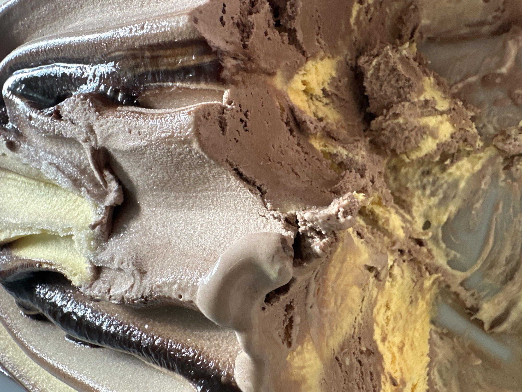 A close-up, top-down, macro shot captures swirls of chocolate and yellow ice cream. The texture is rough and granular in some areas, with smooth, glossy streaks of what appears to be chocolate syrup interspersed throughout. The lighting highlights the different textures and colors, creating a visually rich and appetizing scene of frozen dessert.