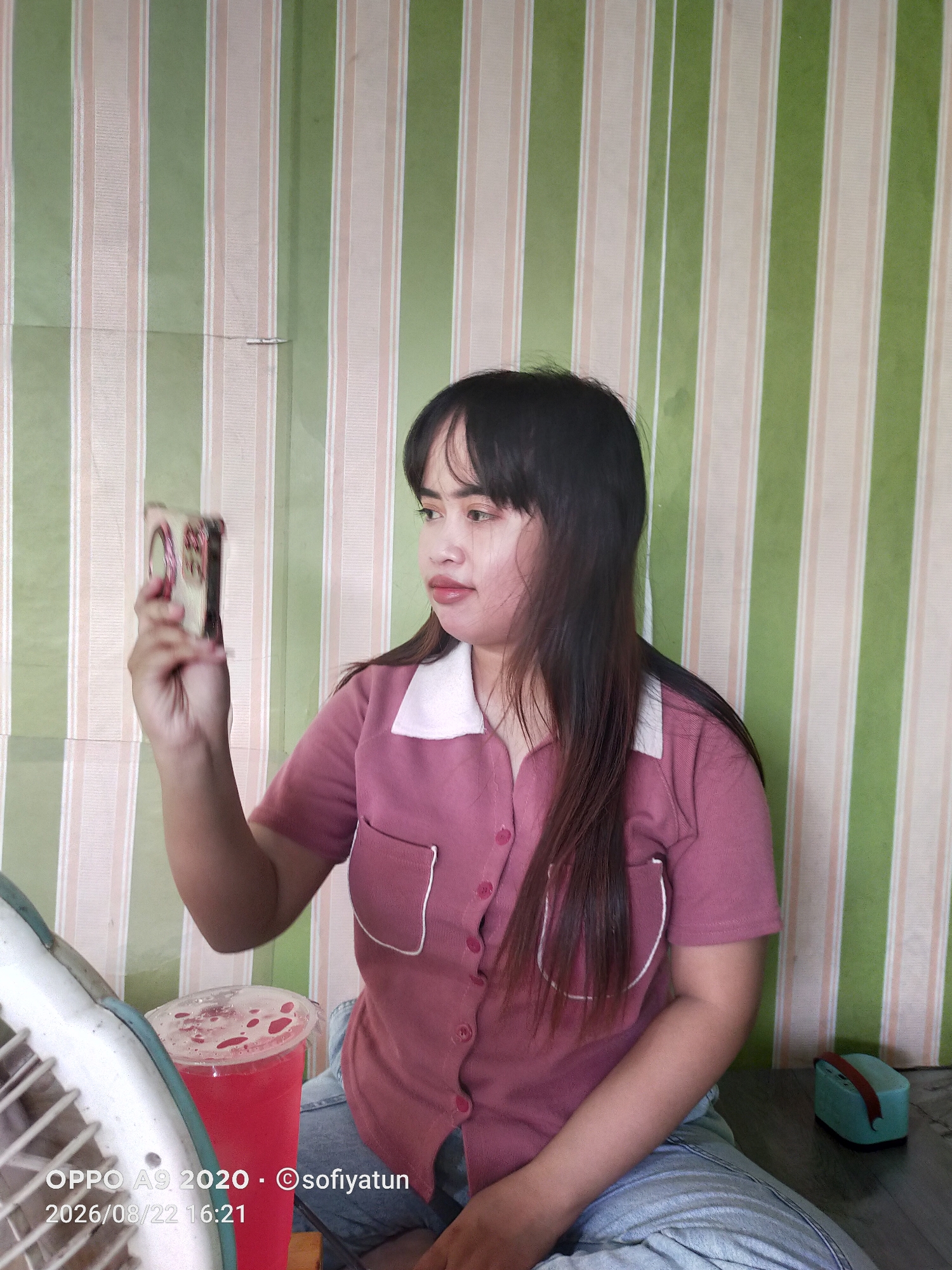 A young woman with long, dark hair and bangs, wearing a pink collared shirt with white trim and light blue jeans, sits indoors. She is holding a small, square object, possibly a phone or compact camera, up to her face as if taking a selfie or looking at it. To her left, a large fan is partially visible, and a red beverage in a clear plastic cup with a lid sits on a table. Behind her, a wall is covered with alternating vertical stripes of light green and beige. On the floor to her right is a small, teal-colored rectangular object with a brown strap. The image is dated 2026/08/22 16:21. This scene is likely in Tayu, Indonesia.