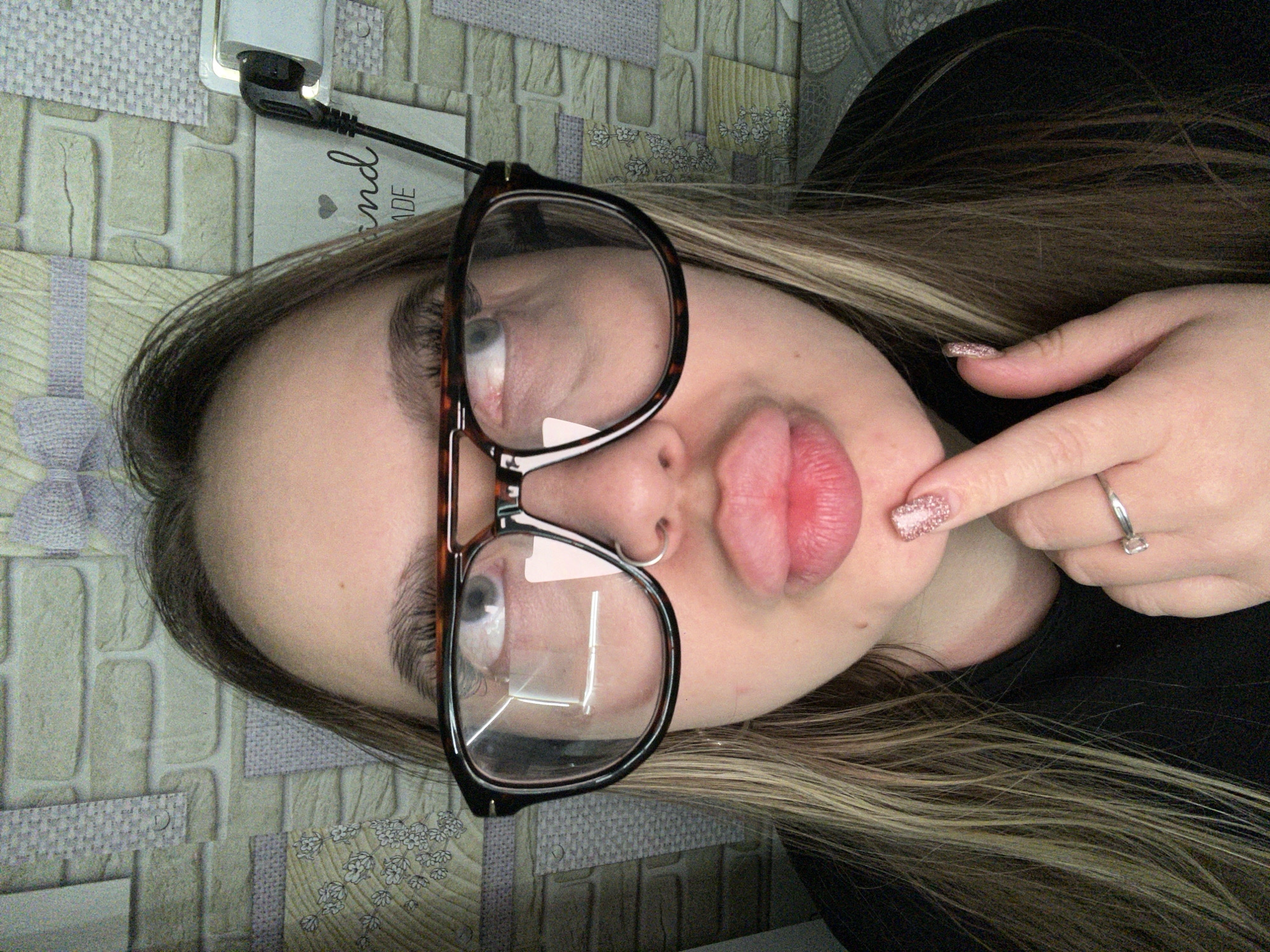 A Caucasian woman with light brown and blonde hair and wearing glasses is lying on a patterned surface. Her lips are pursed. She has a nose ring in her left nostril. Her right hand is raised, with her index finger pointing towards her chin. She is wearing a silver ring with a rectangular stone on her right ring finger. Her fingernails are manicured with a glittery polish. To her left, a white electrical adapter and cable are visible against the patterned surface. The setting appears to be indoors. The location is unknown.