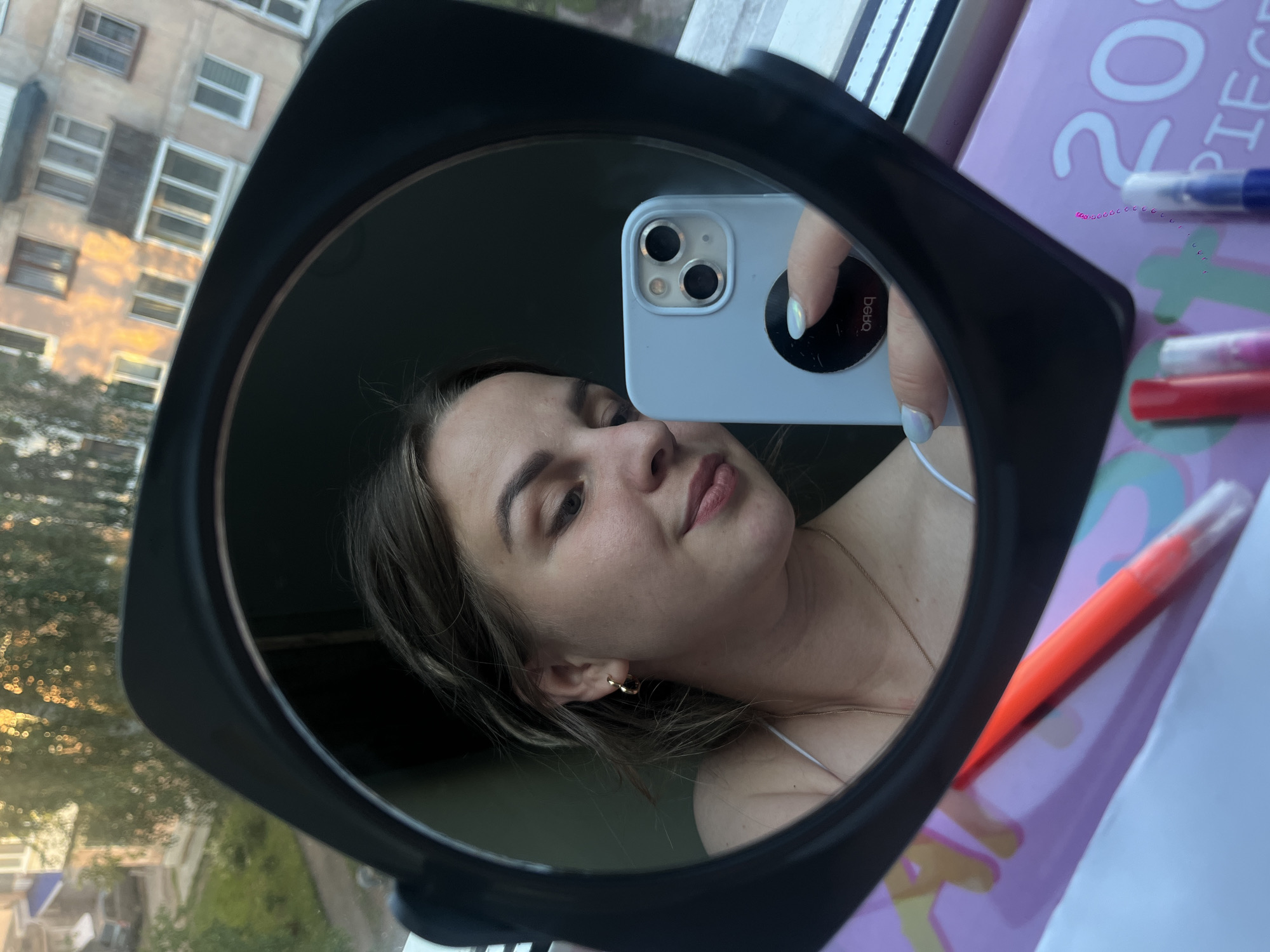 A young woman is depicted taking a mirror selfie, reflected in a black-framed, circular mirror. She holds a light blue smartphone with dual cameras, featuring a white pop socket on the back that clearly displays the text "bebo." The woman has light brown hair, a neutral expression, and is wearing a delicate necklace and a gold-toned hoop earring. Her fingernails are painted with a subtle, pearlescent polish.
The mirror captures not only the woman but also parts of her environment. To the left of her reflection, an outdoor scene is visible: a multi-story residential building with numerous rectangular windows, characteristic of older urban architecture. The building is bathed in the warm, golden light of what appears to be late afternoon or early evening, suggesting clear weather. Green tree foliage is also discernible. This architectural style is consistent with buildings found in cities like Izhevsk, Russia.
To the right within the mirror's reflection, an indoor setting is shown. A light pink box or package is visible, printed with the text "20 PIECES," suggesting art or craft supplies. Beside it, parts of red and blue markers or pens can be seen. The overall scene indicates the woman is indoors, likely near a window, using the mirror to capture both herself and elements of her surroundings.