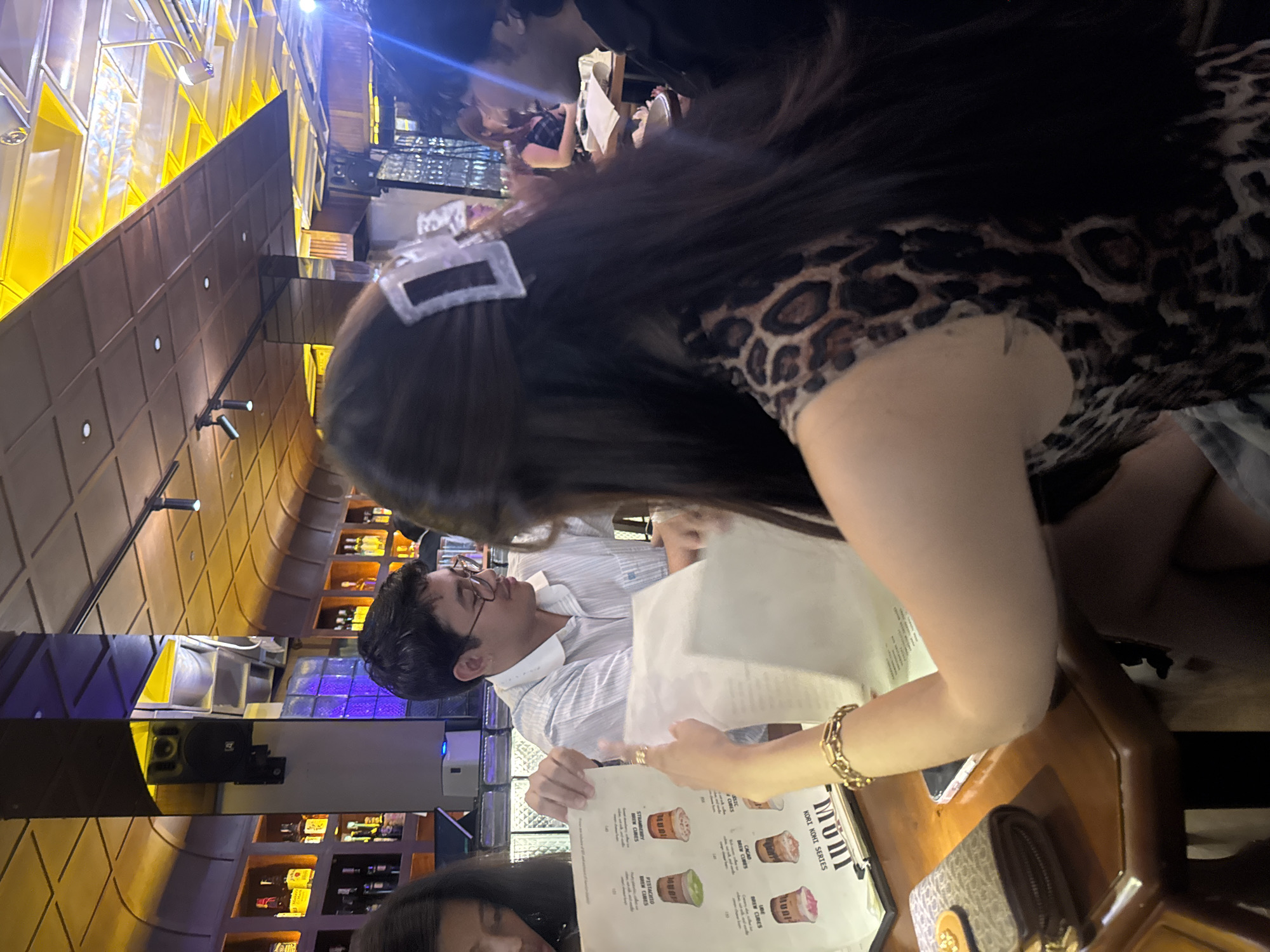 In this image, a group of individuals is seated at a table, possibly in a restaurant or bar. A woman in the foreground, facing away from the camera, wears a leopard-print top and a gold bracelet, and appears to be pointing at a menu. To her left, a man in a striped shirt and glasses is looking at a menu, and a woman to his left is also looking at a menu. On the table, there is a wallet, a smartphone, and menus. The background shows shelves stocked with bottles, characteristic of a bar. The overall lighting suggests an indoor evening setting. The presence of specific bottle brands in the background, such as Johnnie Walker, suggests a potentially upscale establishment. While the exact location is not explicitly stated, the mention of "Davao City, Philippines" in the prompt implies this scene is situated there.