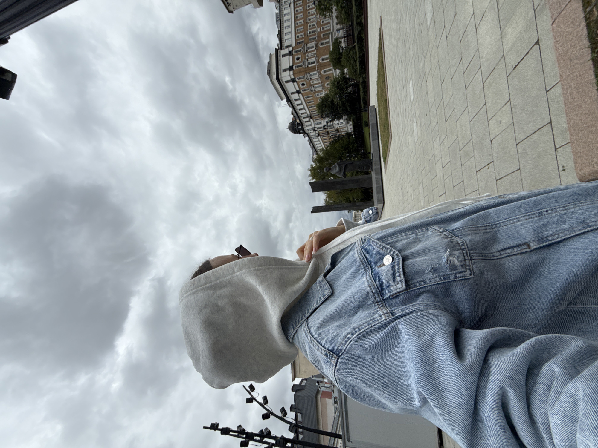 An individual wearing a light blue denim jacket over a grey hooded sweatshirt stands in a city square, looking to their right. The individual's face is partially obscured by the hood and their sunglasses. In the background, there is a multi-story building with a distinct architectural style. To the left, a lamppost with multiple lights is visible, along with the corner of a white structure. In the center of the square, a statue stands on a pedestal, flanked by two dark rectangular pillars. The ground is paved with light-colored square tiles. The sky is overcast with grey clouds. This scene is likely from Moscow, Russia, given the architectural style of the building and the presence of public art.