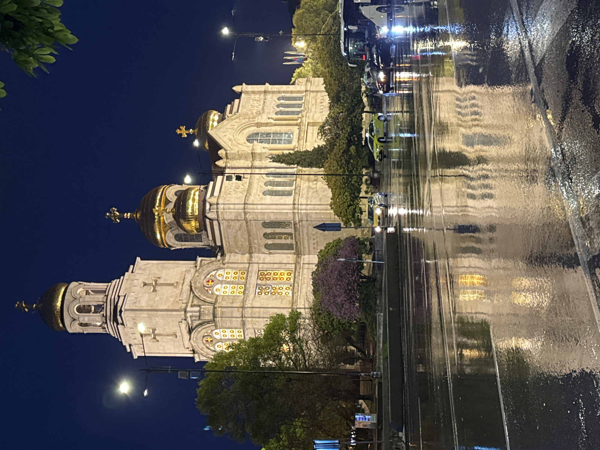An evening street scene in Varna, Bulgaria, captures the illuminated Dormition of the Theotokos Cathedral reflected on a wet road after rain. The time of day is dark night or late evening, indicated by the deep blue sky and artificial lighting.
The central subject is the grand Dormition of the Theotokos Cathedral, a prominent Orthodox church. Its light-colored stone facade is brightly lit, highlighting its intricate architectural details, including multiple arched windows, some of which appear to be stained glass or illuminated from within. The cathedral features several golden domes, each topped with a cross, gleaming under the artificial lights. One large bell tower with an onion dome is visible on the left side of the cathedral.
In the foreground, a dark asphalt road runs parallel to the cathedral. The road is heavily wet, creating a strong, mirror-like reflection of the cathedral's facade, golden domes, and the bright vehicle lights. Several cars, including a distinct light green vehicle, are visible on the road, with their headlights and taillights creating streaking reflections. Other vehicles, possibly a bus, are also present.
Trees and foliage line the street between the cathedral and the road, with some displaying purple blossoms. Additional green foliage is visible in the top left corner. Streetlights are illuminated, adding to the bright reflections on the soaked pavement. No people or visible text are discernible in the image. The overall atmosphere conveys a serene yet vibrant urban nightscape, defined by the interplay of light, water, and architecture.