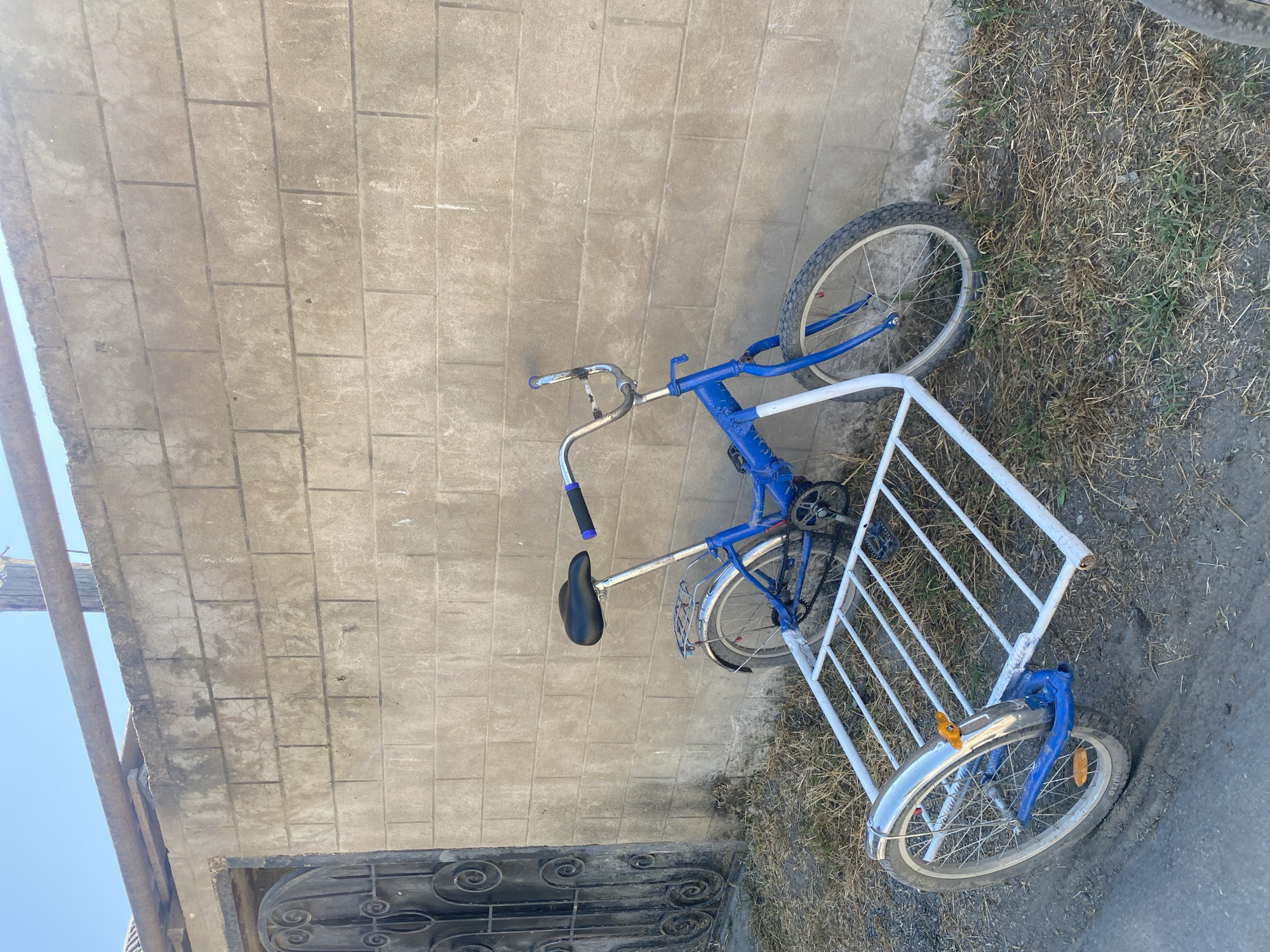A blue and white bicycle with a trailer is parked leaning against a concrete block wall. The bicycle has a black seat and silver handlebars. The trailer is white with silver fenders and a silver wheel. The wall is made of light-colored concrete blocks and has some discoloration. To the left of the wall is a dark metal gate with intricate scrollwork. The ground in front of the wall is covered in dry grass and dirt. The scene appears to be in an outdoor setting, possibly a yard or alley. The image does not provide enough information to definitively place it in the city of Samur, Russia.