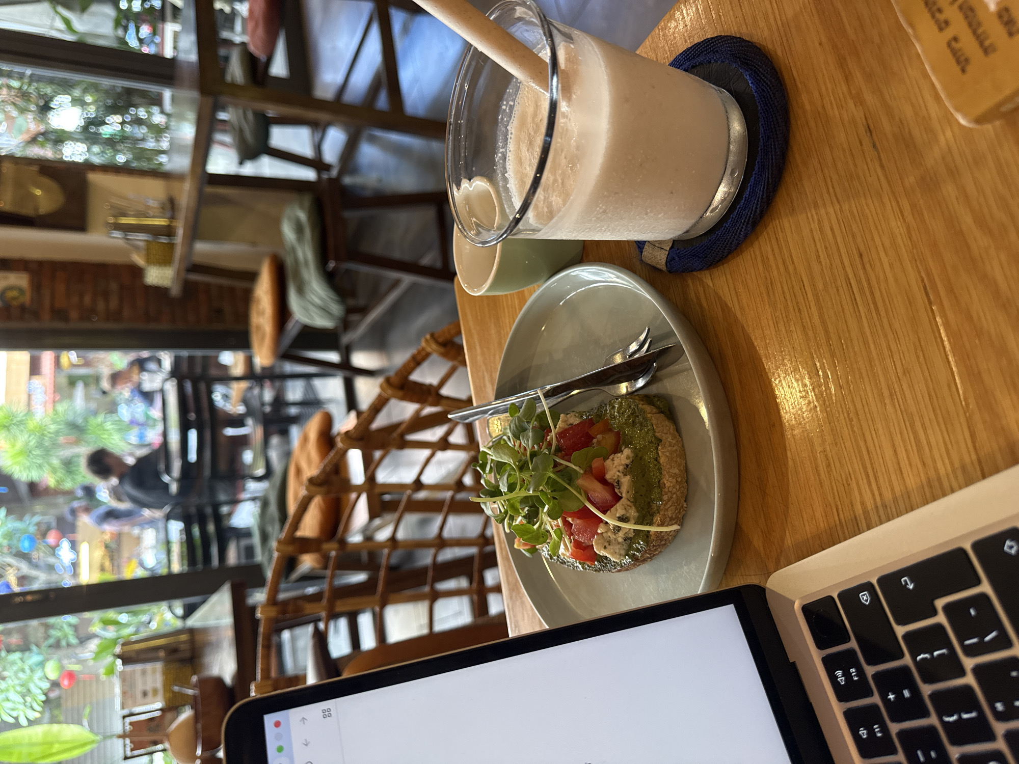 On a wooden table in a café in the city of Unknown, Unknown, lies a laptop computer, a plate of bruschetta, and a glass of a smoothie-type beverage with a straw. The laptop is open, displaying a white screen, and the keyboard is visible on the right side. The plate contains a piece of bruschetta topped with pesto, tomatoes, cheese, and greens. A fork and knife rest on the plate next to the bruschetta. A glass of smoothie is on a blue coaster. The café appears to be busy with patrons sitting at tables in the background, visible through a large window. The interior of the café has wooden furniture, including high chairs with woven backs.