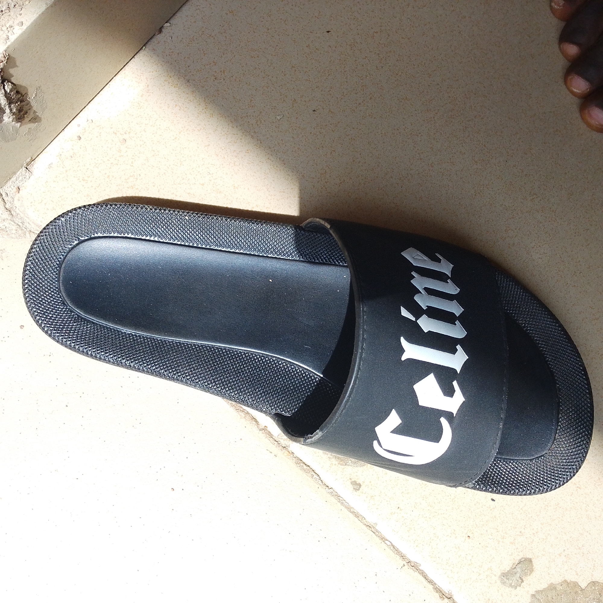 This media file shows a black slide sandal with the word "Celine" in white, gothic-style lettering on the strap. The sandal is placed on a tiled surface, possibly a floor or patio. The tiles are light beige with darker speckles. A shadow from what appears to be an overhead object or structure falls diagonally across the scene, suggesting it is outdoors during the daytime. The lighting is bright, with a harsh shadow, indicating sunny weather. In the upper right corner of the image, part of a person's foot, specifically toes with dark skin and light nails, is visible, suggesting the sandal might be in use or about to be used. The overall scene is simple, focusing on the sandal and its immediate surroundings. There is no discernible emotional content, and the location is not specified beyond the tiled surface.