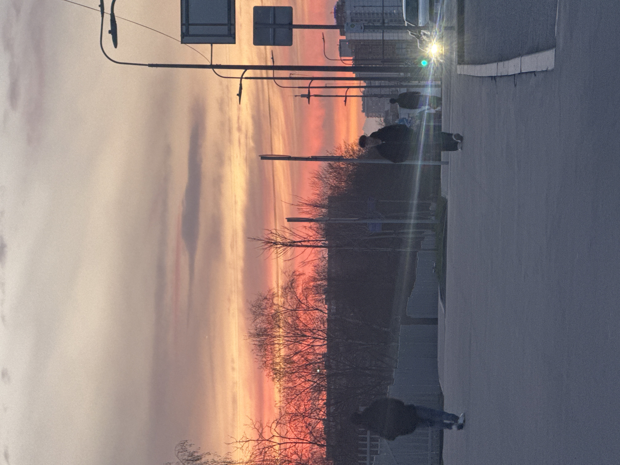 The image depicts a street scene at sunset in what appears to be an urban area, possibly Moscow, Russia given the architecture. The sky is a mix of orange, pink, and gray hues. Silhouetted against the sky are bare trees. In the foreground, there is a paved road with pedestrians walking along the sidewalk. There are street lights and traffic signals visible, and a car with its headlights on. Buildings and a green traffic light are visible in the distance. The scene suggests a late evening or early morning atmosphere in a city environment.