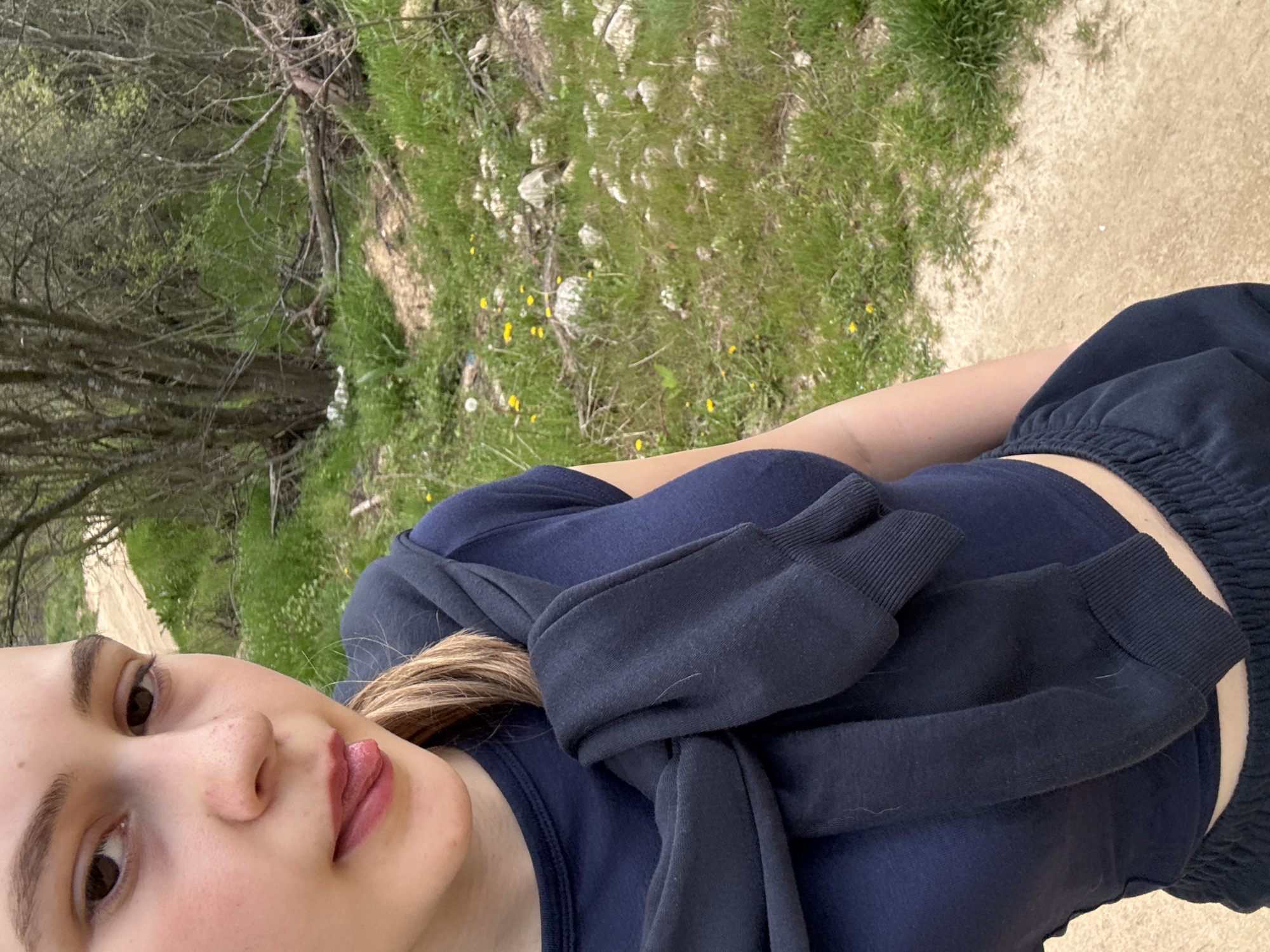 A selfie of a young woman outdoors in daylight. She has light skin, brown eyes, and blonde hair. She is wearing a dark blue crop top and dark blue sweatpants. A dark blue sweatshirt is draped around her shoulders. The background includes trees, grass, rocks, and a dirt path. The location is not identified in the city of Panagyurishte, Bulgaria.