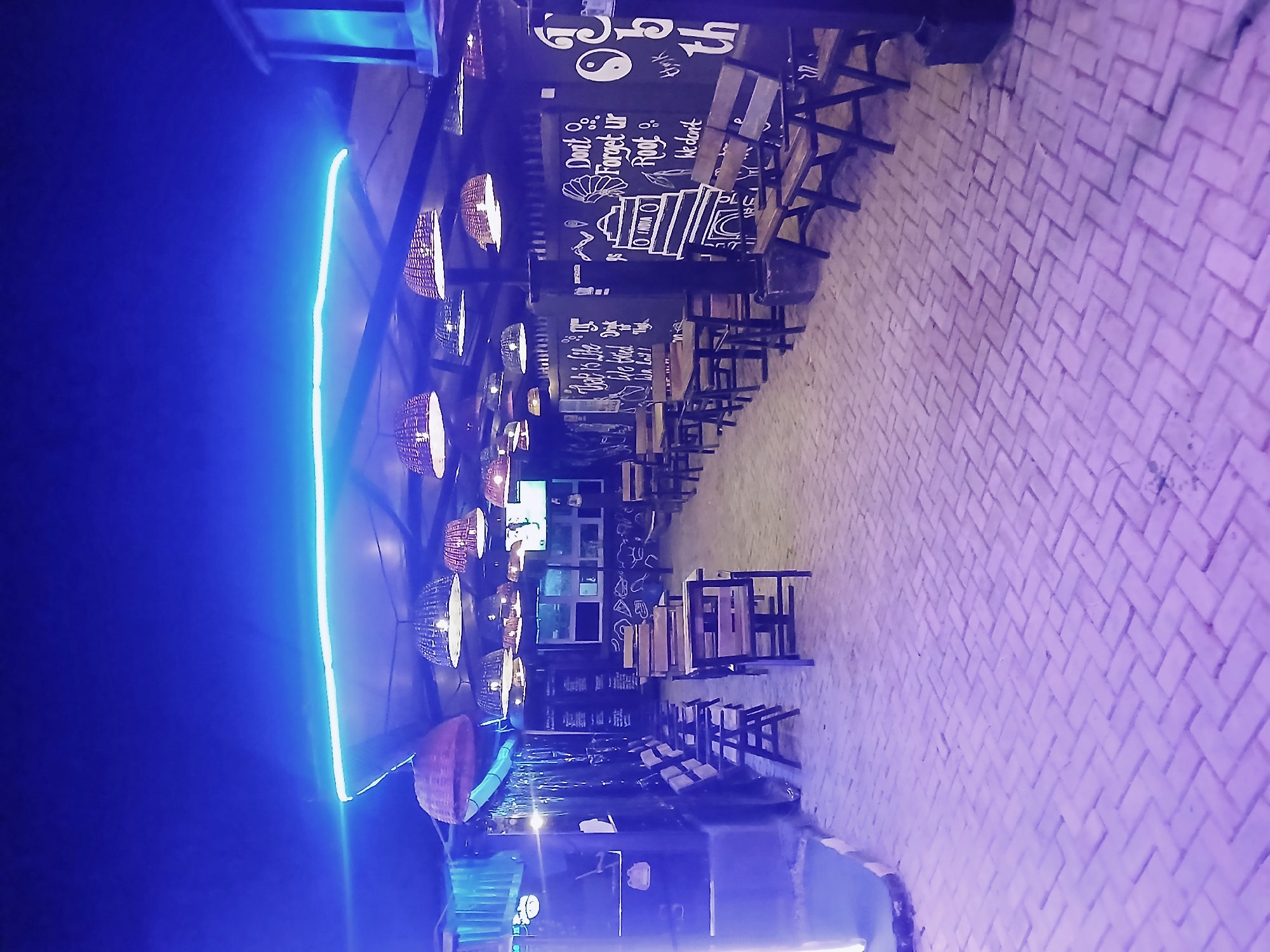 A low-angle, rotated outdoor scene at night in an unknown city shows a bricked walkway lined with dark wooden tables and chairs. The area is illuminated by a string of round wicker-like lamps hanging from the ceiling and a neon blue light strip running along the side. The ceiling and lamps are on the left, the brick walkway and chairs on the right. A black wall with white writings, including the words "Don't Forget Ur Root," runs along the length of the walkway behind the chairs. The sky is dark blue. A TV screen is visible between the lamps.