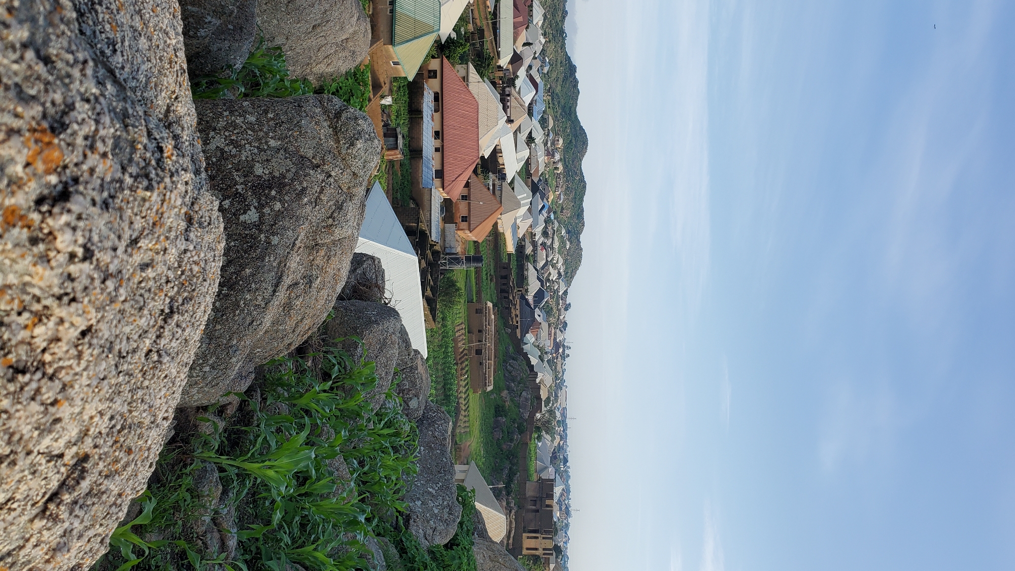 The image depicts a residential area in the city of Jos, Nigeria, as seen from a rocky, elevated vantage point. In the foreground, large, grey boulders covered in lichen dominate the view, with a patch of green vegetation growing between them. Beyond the rocks, the mid-ground reveals a sprawling settlement of houses with various colored roofs, many appearing to be constructed with corrugated metal. Some houses are well-maintained, while others show signs of ongoing construction, with exposed brickwork and incomplete structures. A water tank on a stand is visible near one of the houses. The background is characterized by rolling hills and a hazy sky, suggesting a moderately clear day. The overall impression is of a developing urban landscape situated amidst natural rocky terrain. There are no people or animals visible in the frame.