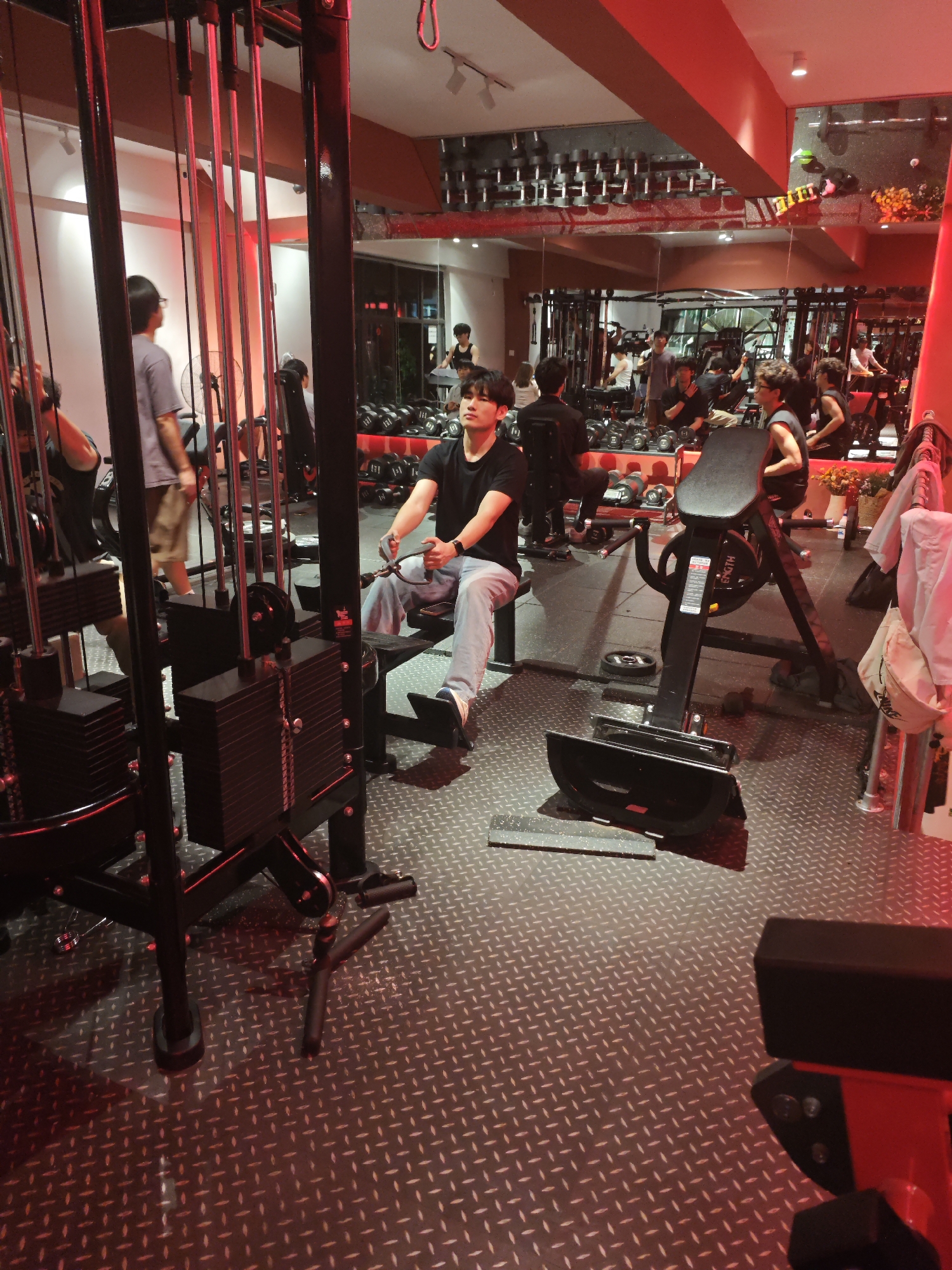 The image depicts the interior of a gym in Chongqing High-tech Zone, China. Several people are engaged in exercise activities around weightlifting equipment. A man in a black t-shirt and light-wash jeans is seated on a rowing machine and appears to be in the middle of a workout. The gym has a mirror along one wall that reflects the activity and equipment. Dumbbells are neatly arranged on racks behind the exercisers. The space is lit with red lighting. The floor has a textured, non-slip surface. Weight machines, benches, and other exercise gear are visible throughout the space. The gym appears to be active with multiple individuals using the equipment.