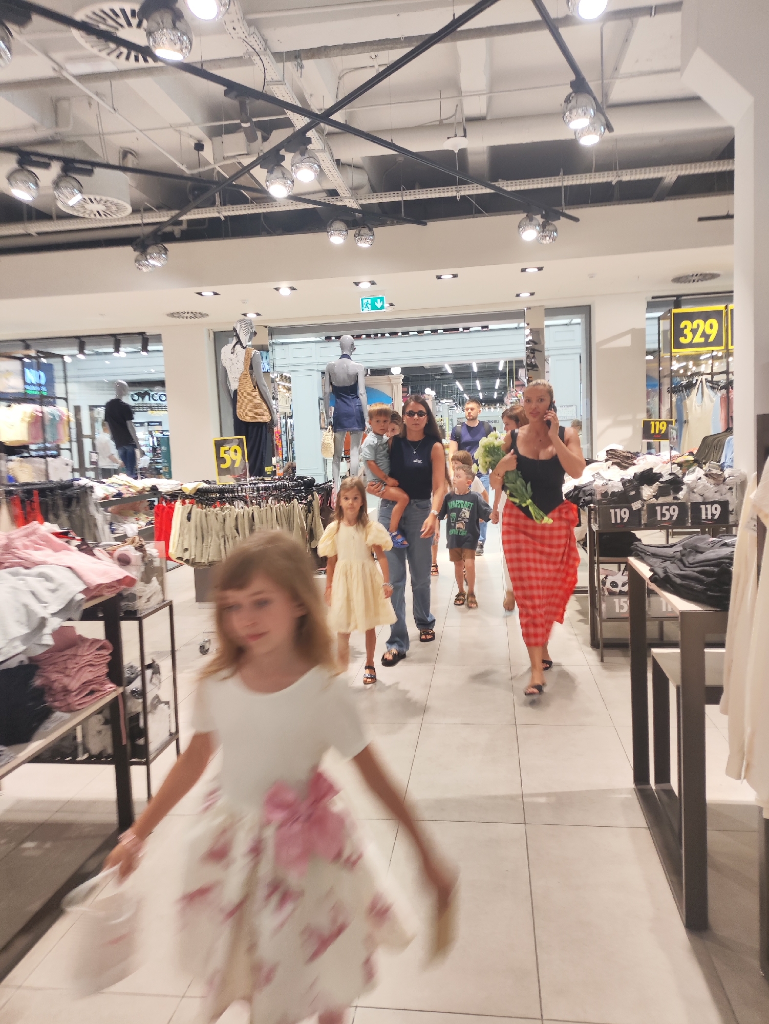 The image depicts a group of people walking through a retail store, likely a clothing store, in Chișinău, Moldova. In the foreground, a young girl in a white dress with a large pink bow is blurred due to motion. Behind her, a woman in a black top and a red plaid skirt is talking on her phone, holding a bouquet of flowers. To her left, a woman in a dark top and jeans is carrying a child. Other children and adults are visible walking in the same direction. The store environment features clothing racks, displays of garments, and overhead lighting. Signage indicating prices and possibly department numbers, such as "329" and "119," is visible. The flooring appears to be tiled. The overall scene suggests a shopping trip.