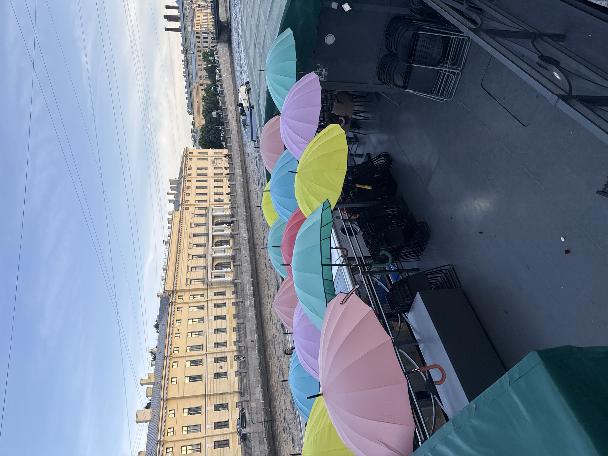 The image shows a section of the Fontanka River in Saint Petersburg, Russia, on a cloudy day. In the foreground, a boat is docked, covered with an array of colorful umbrellas, including pink, yellow, blue, green, and purple. Stacked black chairs are visible on the deck of the boat, some underneath the umbrellas and others to the side. A dark grey storage box with an orange handle is placed on the deck. In the background, along the riverbank, a long, pale yellow building with numerous windows and classical architectural elements stands. Trees line the embankment between the building and the river. Further in the distance, more buildings and industrial chimneys are visible. A white boat with a few people on board is navigating the river. The sky is a pale blue with some wispy clouds and visible power lines stretching across it.