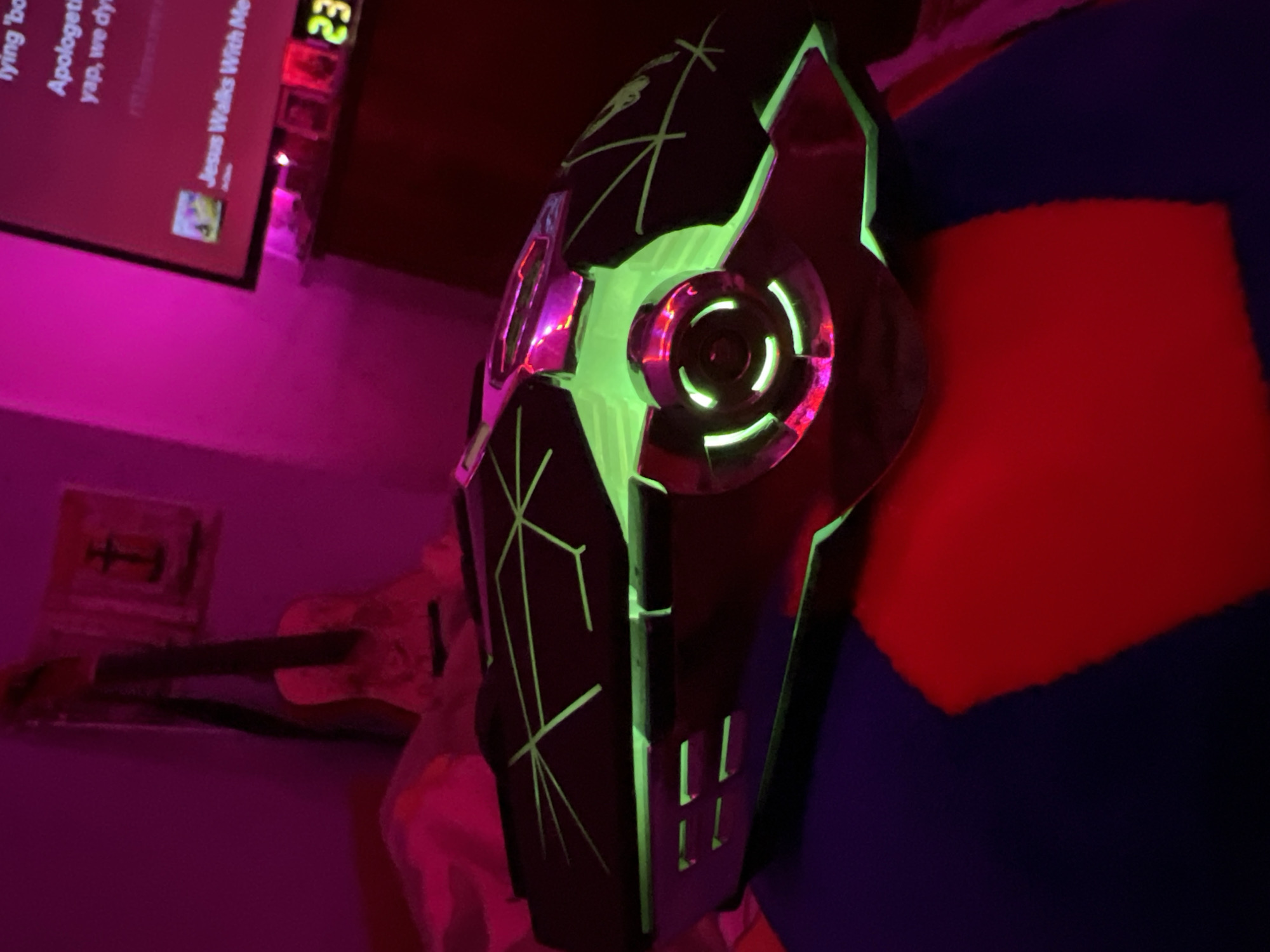 A dark interior space, identified as a room in Debelt, Bulgaria, is illuminated primarily by artificial magenta light, casting a strong hue on the walls and ambient environment. In the foreground, a dark-colored, angular electronic device with integrated green and magenta illumination is positioned on a textile surface composed of red and dark blue sections. The device features geometric patterns rendered in green light on its upper surface, along with bright green internal lighting visible through vents and a central, circular mechanism that also emits or reflects magenta light. To the left, an acoustic guitar with a light-colored body and dark neck leans vertically against a magenta-illuminated wall. To the right, a flat-screen television displays text in magenta characters, including fragmented phrases such as "lying 'bo", "Apologet", "yap, we dy", and clearly legible "Jesus Walks With Me". Below the television, a digital clock displays the green numerals "23". No individuals are visible within the frame.
