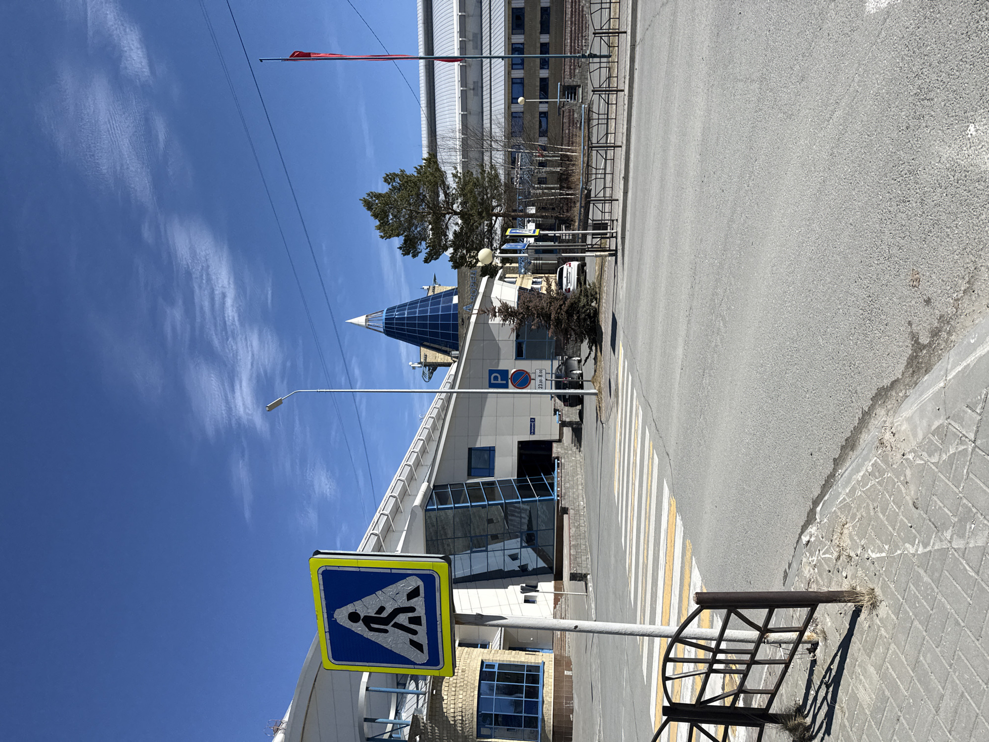 The image depicts a street scene in Beloyarsky, Russia. A crosswalk sign is prominent in the foreground, with a pedestrian crossing extending across the road. A building with a pointed, glass-covered roof is behind the sign. A street light is positioned nearby, reaching upwards to the sky. The sky is clear blue with wisps of clouds. A parking sign is visible near the building. Further in the background, a multi-story building is visible behind a tree. A red flag is visible near the top of the multi-story building. The street is paved with asphalt and a metal barrier is present on the side of the road.