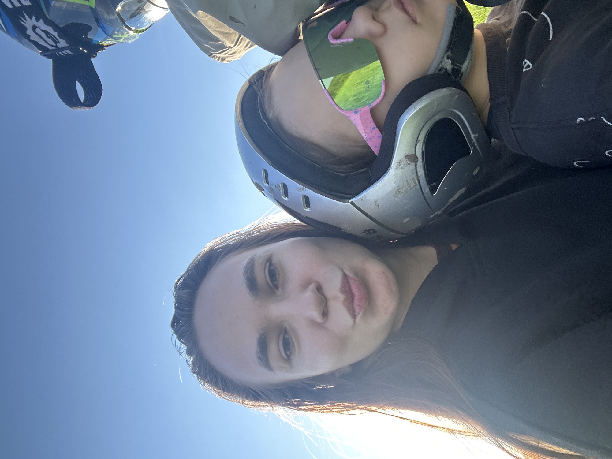 This image depicts a close-up, selfie-style portrait of two individuals, with a third partially visible, set against a clear blue sky in the city of Kharnavy, Russia.
The foreground features an adult female on the left. She has long, dark brown hair, illuminated by sunlight from the left, and is looking directly at the camera with a puckered lip expression. She wears a dark-colored hooded garment, likely black, with a glimpse of a reddish-brown undergarment visible at the neck.
To her right and slightly behind her is a younger individual, likely a child. This individual wears a scuffed, silver/grey full-face helmet with dark interior padding. Oversized, frameless-style sunglasses with reflective green lenses and speckled pink frames obscure the eyes. The lower part of the child's face is visible beneath the sunglasses, with lips appearing relaxed. The child wears a dark-colored long-sleeved shirt, possibly black, with white graphic markings visible on the right shoulder and sleeve. The child's head is tilted slightly down and right, resting in close proximity to the adult female.
In the upper right background, a portion of a third individual is visible, identifiable by a helmet featuring blue, grey, and black patterns with white graphic elements. A black goggle strap is visible across this helmet. Below this, a light-colored fabric garment is discernable.
The background consists solely of a clear, bright blue sky, indicating an outdoor setting with strong, clear lighting, consistent with either early morning or late afternoon sun originating from the left. No discernible ground features, buildings, or additional environmental elements are present. The presence of multiple helmets suggests an activity such as motorcycling, dirt biking, or ATV riding.