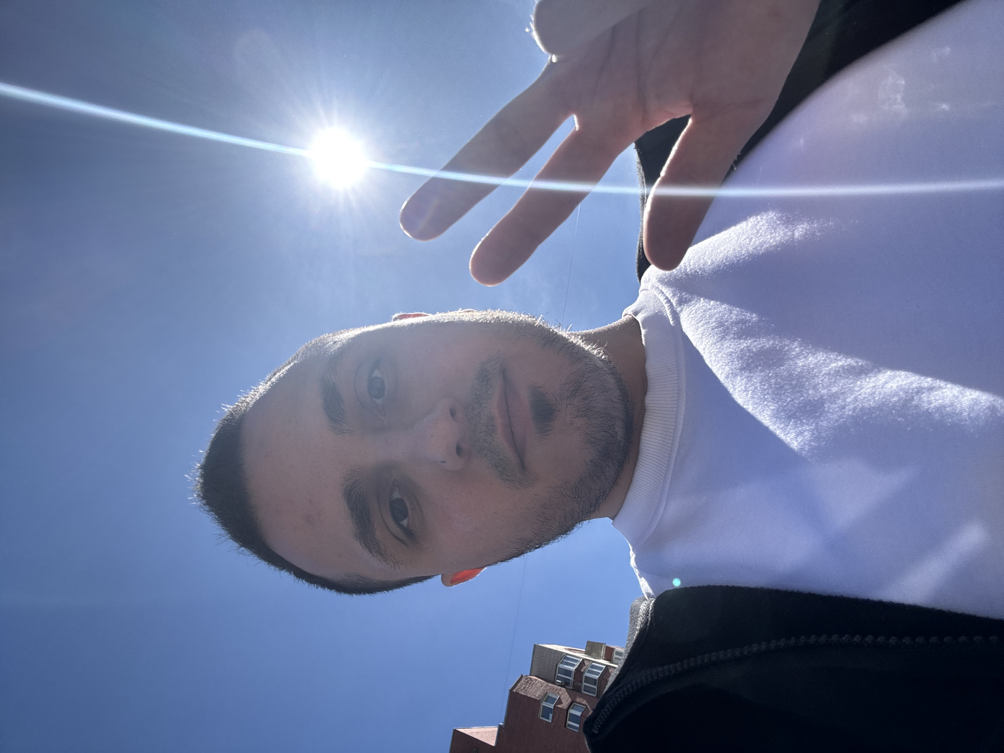 A young man is taking a selfie outdoors with a bright sun in the background. He is wearing a white crew neck sweatshirt and a black jacket. His right hand is raised, with two fingers extended in a peace sign. In the background, there is a blue sky and a brick building. The image is captured in a close-up perspective, focusing on the man's face and upper body.