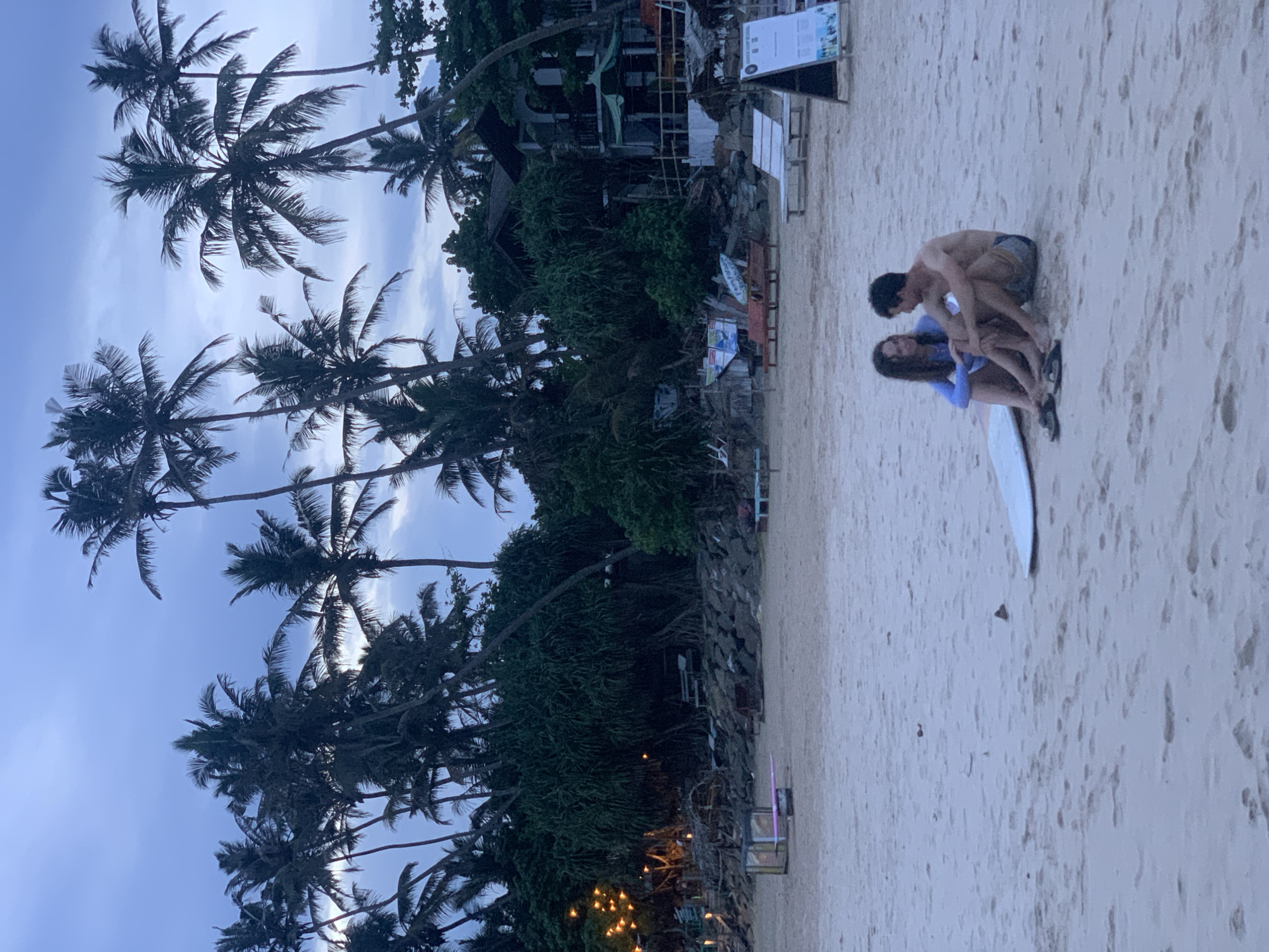 The image depicts a beach scene in Kathaluwa, Sri Lanka, with palm trees lining the left side of the frame. A couple sits on the sand, near a surfboard, with the man embracing the woman. Vegetation and structures are visible behind them. The sky is overcast.