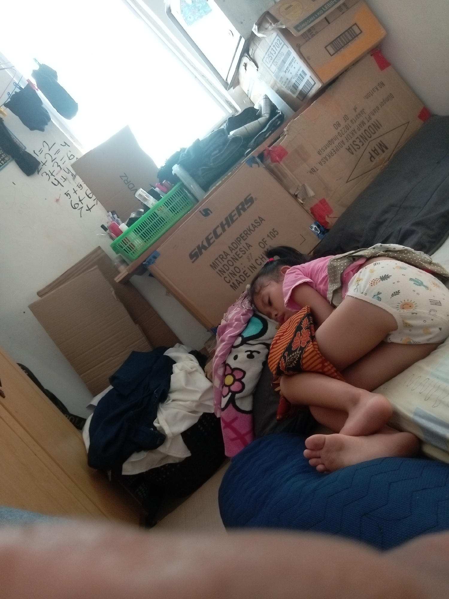 This media file shows a young child sleeping peacefully on a bed, partially covered by blankets. The child is wearing a pink t-shirt and a diaper adorned with cartoon patterns. Next to the child is a pillow with a Hello Kitty design. The surrounding environment appears to be a bedroom or living area with several cardboard boxes stacked, one of which is labeled "SKECHERS" and another with "MAP INDONESIA". Other objects visible include clothes in a basket, a green plastic container, and some writing on a white wall that seems to be math equations. The lighting suggests it might be daytime, possibly a nap time. The presence of multiple cardboard boxes hints at a move, recent delivery, or storage. The overall scene evokes a sense of domesticity and tranquility. The text visible on the boxes provides brand names and origin information, identifying the location as Indonesia.