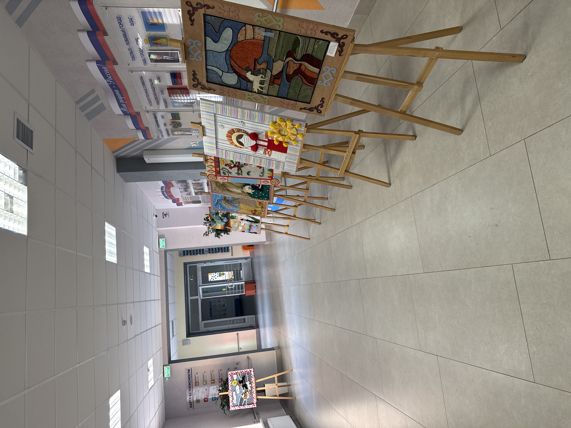 The image shows an indoor hallway with a series of framed artworks displayed on wooden easels. The hall features a tiled floor, a white ceiling with rectangular lighting fixtures, and a glass-enclosed entrance at the far end. Along the upper left wall are decorative banners in the colors of the Russian flag with the text "Моя родина - Россия! (My homeland - Russia!)" and "ТОБОЛЬСК - ЛЮБИМЫЙ ГОРОД! (Tobolsk - beloved city!)" along with depictions of the flag of Tobolsk, Russia, and various images, as well as other texts relating to the Tyumen Oblast and state symbols of the Russian Federation. At the far end of the hall, near the entrance, is a safety information poster on the wall. The location appears to be a gallery or exhibition space, possibly within a public building in Tobolsk, Russia. There are no people visible in the image. The focus is on the display of artworks.