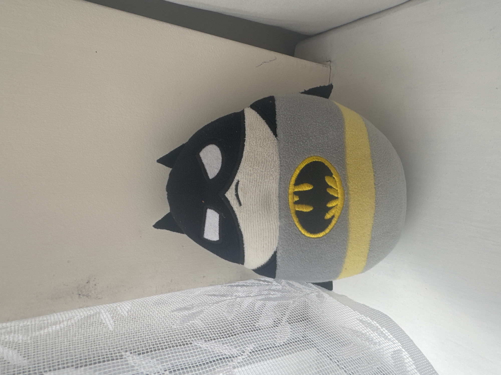 A Batman plush toy leans against a white wall and ceiling in a room. Part of a white, mesh-like object is in the foreground, obscuring the lower portion of the image. The toy has a black and white face and gray body with a yellow stripe featuring the Batman logo. The location in Welshpool, United Kingdom is undetermined.