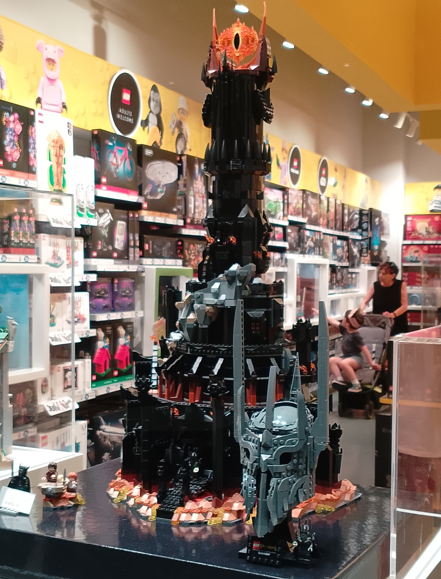 The image shows a large, elaborate Lego build of a dark tower, possibly from a fantasy movie, on display in a store. The tower is black and grey with fiery red accents at its base. In front of the tower, there are Lego minifigures, including what appears to be a dark figure in armor and two smaller figures with reddish-brown hair, possibly hobbits. There is also a close-up of a detailed Lego helmet or mask in the foreground.
In the background, a woman is pushing a stroller with a child. The store shelves behind them are filled with various Lego boxes, suggesting a toy store. The wall behind the shelves is painted yellow and features circular signs with the Lego logo and the text "ADULTS WELCOME." Other Lego figures are visible on the yellow wall, including a pink pig character, a gorilla, and other generic figures. The lighting suggests an indoor, well-lit environment, likely during the day. The overall scene depicts a retail environment with a prominent Lego display. A price tag is visible near the front display, though the price is difficult to read. The Lego set name "SAURON" is visible on a baseplate beneath the helmet.