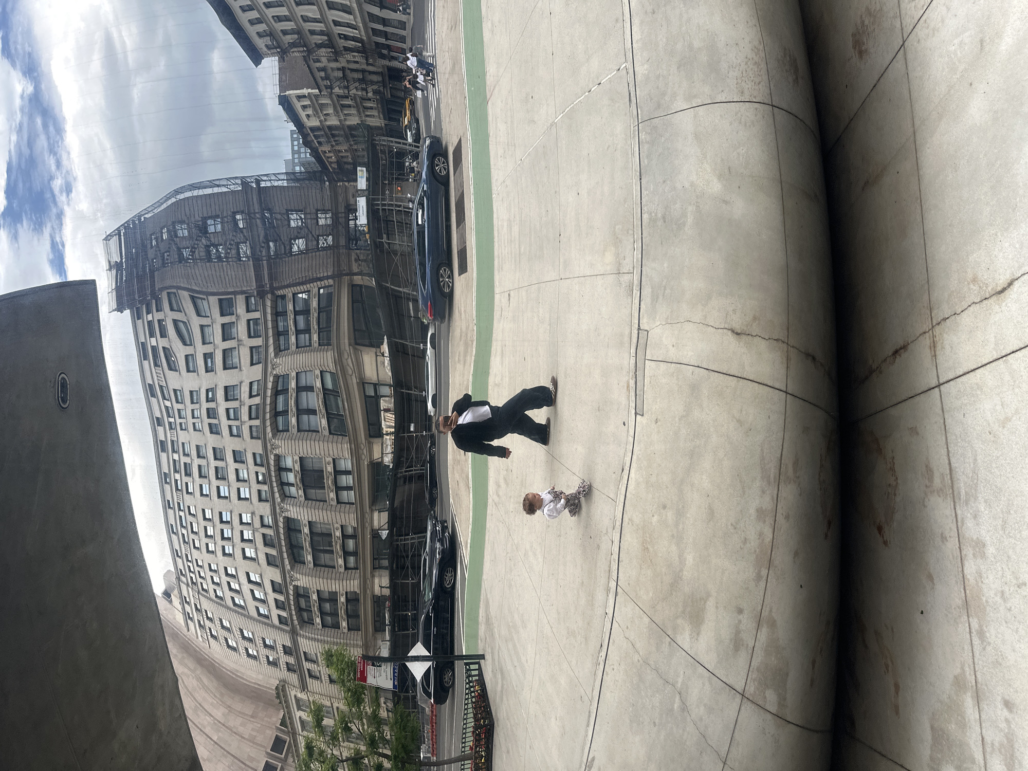 The photograph shows a father and child standing in front of the Cloud Gate sculpture, also known as "The Bean", located in Chicago, Illinois. The reflection in the sculpture captures the city's skyline, including a tall building with multiple windows and scaffolding. A street with cars and a green bike lane is also visible in the reflection. The father is wearing a suit, and the child is wearing a patterned outfit. They are standing on a concrete surface with visible seams. The sculpture itself is made of polished metal, creating a distorted and curved reflection of the surrounding environment.