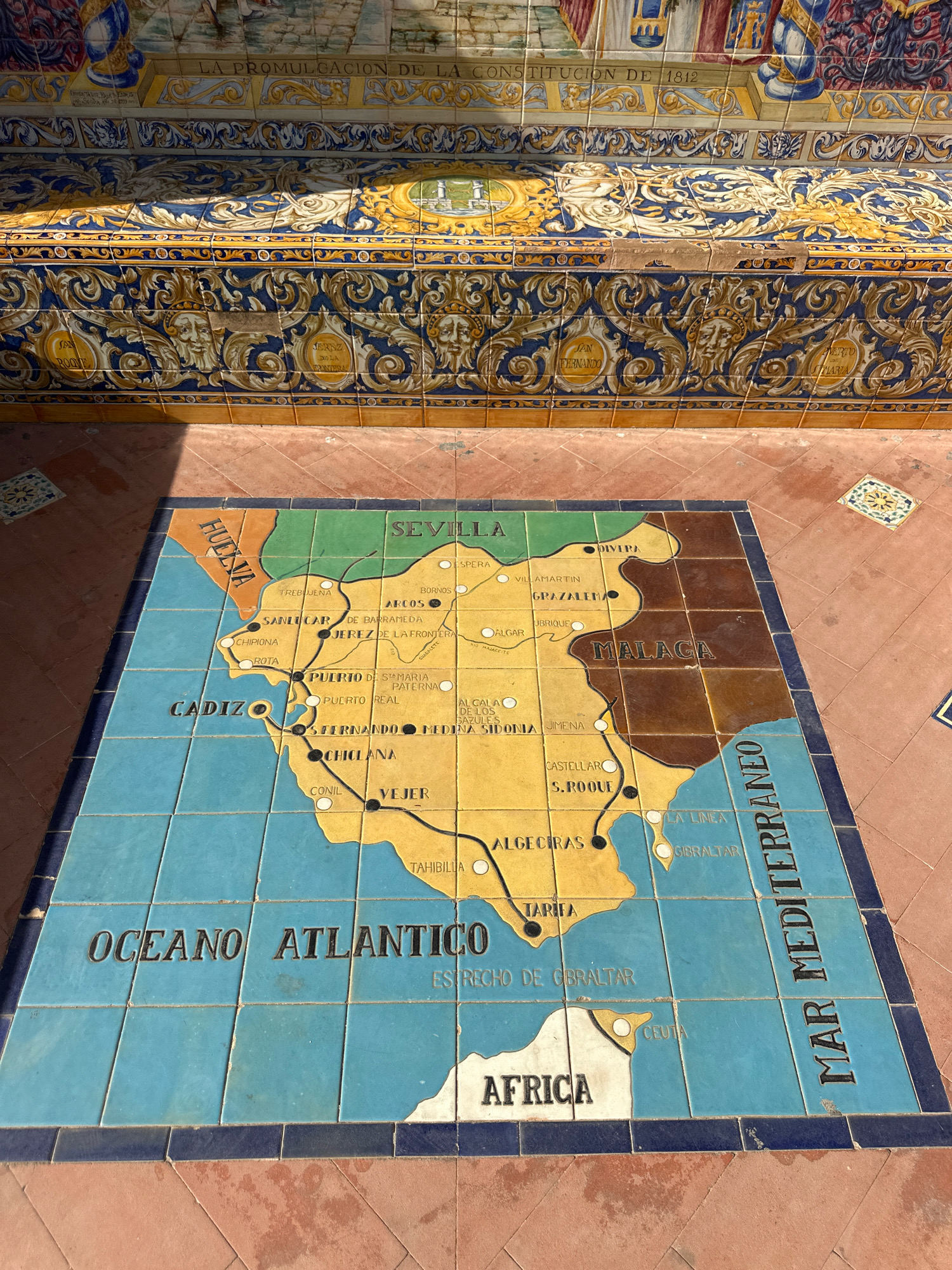 The image shows a detailed tile mosaic map displayed on the ground in Seville, Spain. The map depicts a portion of Andalusia, Spain, including cities like Seville, Cadiz, Jerez de la Frontera, and Malaga. It also shows the Atlantic Ocean and the Mediterranean Sea, with a representation of Africa in the lower right corner. The mosaic is surrounded by a tiled walkway and a decorative tiled wall above. The wall features intricate patterns and inscriptions in Spanish. The mosaic itself is colorful and appears to be well-preserved, though some tiles show wear and tear. The scene is illuminated by natural light, casting shadows on the ground.
