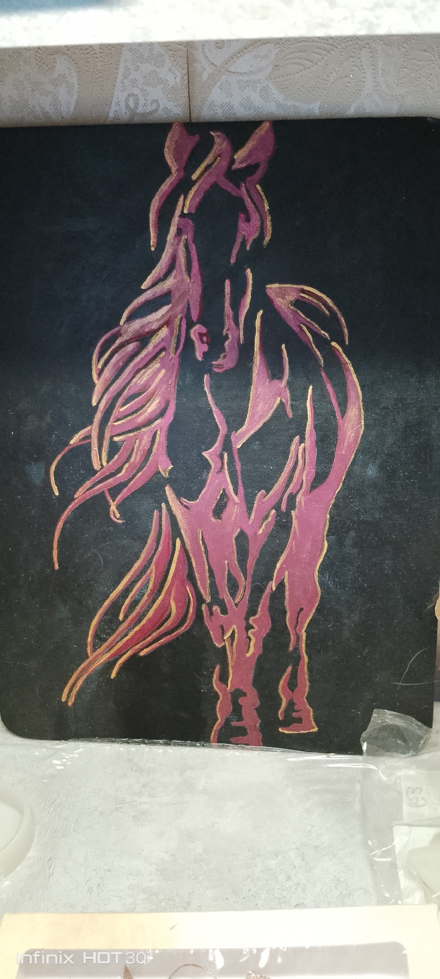 The image is a close-up, vertical shot of a painting of a horse on a dark background. The horse is depicted in a stylized, abstract manner with flowing lines of deep pink and gold paint. The painting appears to be on a canvas or board. The background is a solid, dark color, possibly black. The painting is positioned on a surface that is mostly out of focus, but it appears to be a flat surface with some texture. There is a hint of a white, patterned object in the upper portion of the image, and some clear plastic sheeting is visible in the lower right corner. There is no visible information indicating the location of this image in Kedrovy, Russia.