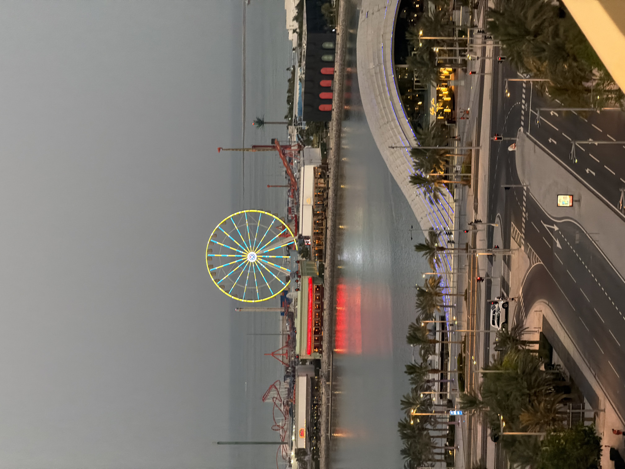An aerial shot captures an amusement park and a modern building complex at dusk. A Ferris wheel with yellow and blue lights is prominently displayed in the center, reflecting on the calm water. Adjacent to it, a roller coaster track curves through the park. The amusement park buildings are illuminated with red and yellow lights. A contemporary building with a curved, illuminated roof is situated in the foreground, with palm trees lining the walkway. Streets with traffic lights and a few cars are visible in the lower portion of the image. The sky is overcast, creating a diffused light that gives the scene a serene, twilight atmosphere.