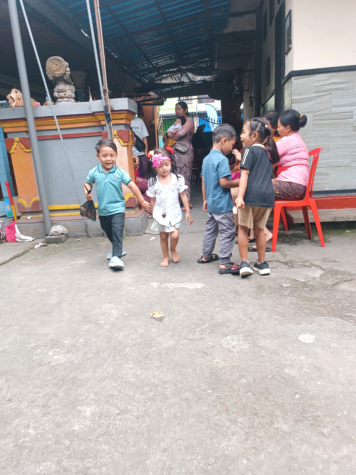 In Tabanan, Indonesia, a group of people, including children and adults, are gathered outdoors. The scene appears to be in a courtyard or alleyway with a concrete ground. Several children are in the foreground, with a boy in a blue polo shirt and dark pants running while holding a dark bag. Next to him, a young girl in a white dress with a flower headband is walking. Behind them, a group of adults and older children are conversing. One adult woman, wearing a pink top, is seated on a red plastic chair, facing away from the camera. Another woman, also in pink, is holding a baby and is partially visible. The background shows a building with an overhang roof supported by columns. Decorative elements, possibly part of a shrine or temple, are visible on the left side of the image. There are also some discarded items on the ground, including a pink cloth and a piece of paper. The overall atmosphere suggests a casual gathering or a community event.