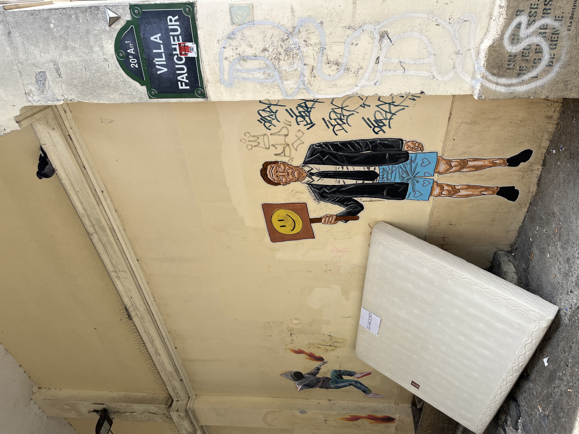 An outdoor scene in Paris, France, features a light yellow wall adorned with various forms of graffiti and street art. To the right, a street sign indicates "20e Arr. Villa Faucheur," marking the location. A pigeon is perched on a ledge above the sign. On the wall, a mural depicts a man in a suit jacket and shorts, holding a sign with a yellow smiley face. To the left of this mural, another piece of street art shows a hooded figure with flames in its hands, with smaller flame graphics on the wall nearby. In front of the wall, a large white mattress rests against it. The ground is littered with debris.