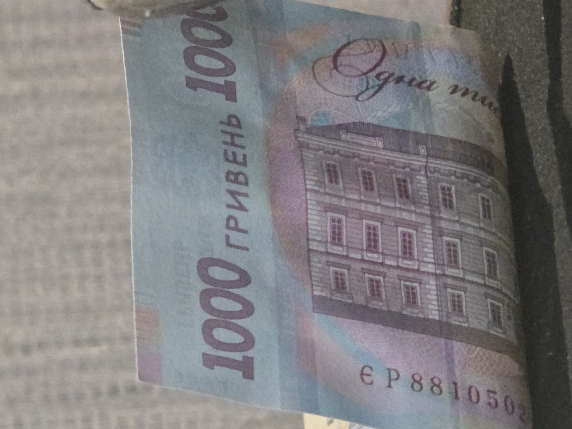 The image shows a close-up of a Ukrainian 1000 hryvnia banknote. The banknote is predominantly light blue and features a building on the lower half. The number "1000" is prominently displayed in purple on the left and right sides of the banknote. Text in Ukrainian, including "ГРИВЕНЬ" (hryven'), is also visible. A serial number, "EP8810502", is printed vertically along the left edge of the banknote. A stylized signature or symbol is present on the right side. The banknote appears to be slightly creased and is resting on a dark, textured surface. The background is a blurred, grayish, textured material. No specific location in a city is identifiable from the image.