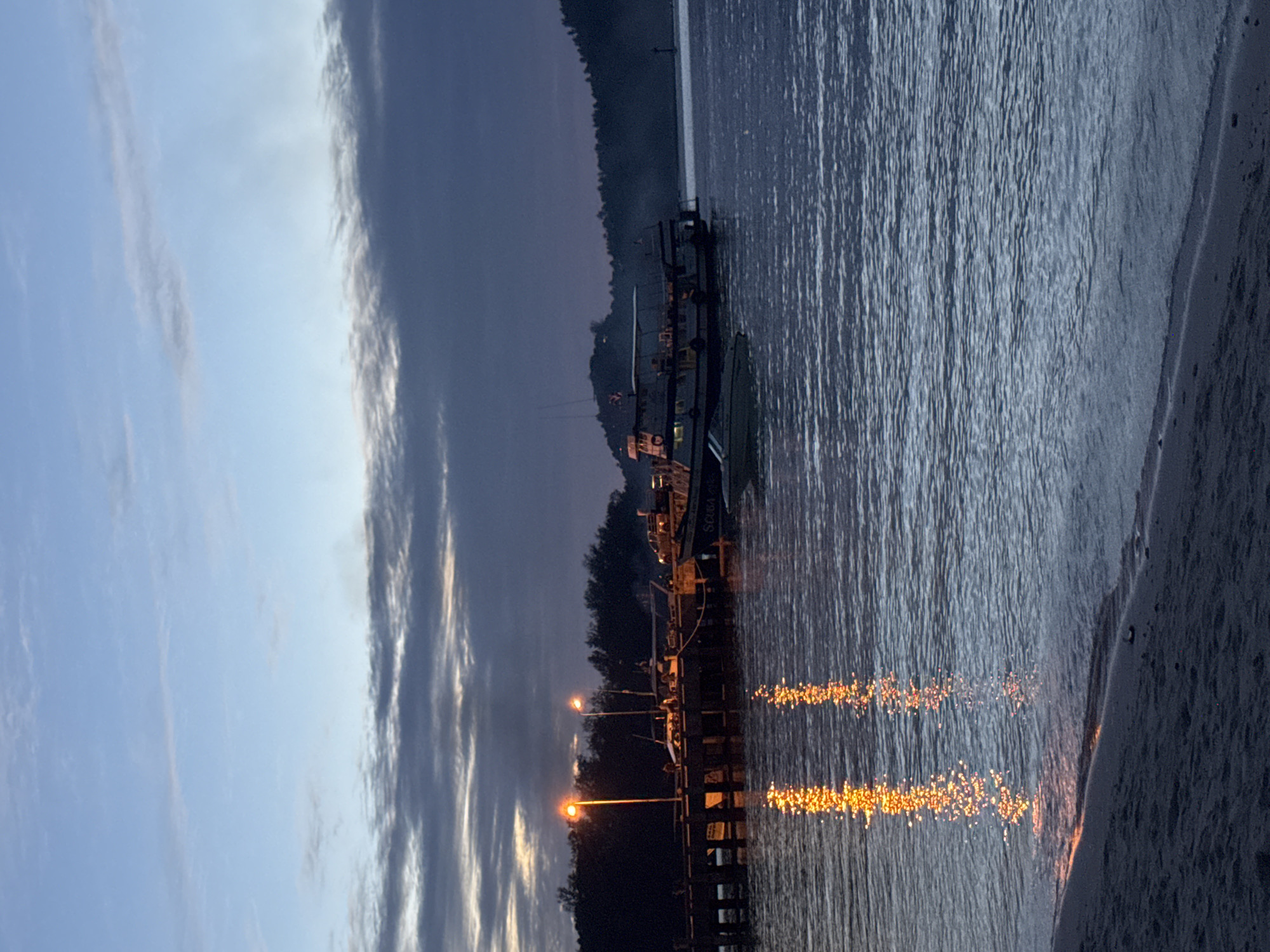 This image depicts a waterfront scene at dusk in Koh Phangan, Thailand. A wooden pier extends into the water, lined with several boats. Two orange streetlights illuminate the pier, casting reflections on the calm sea. The sky is a gradient of blues and grays, with dark clouds gathering on the horizon. In the background, a forested hill rises, and on the right side of the frame, another boat with the text "SCUBA" is visible. The foreground shows a sandy beach leading to the water's edge. The overall atmosphere is peaceful and serene, capturing the transition from day to night.