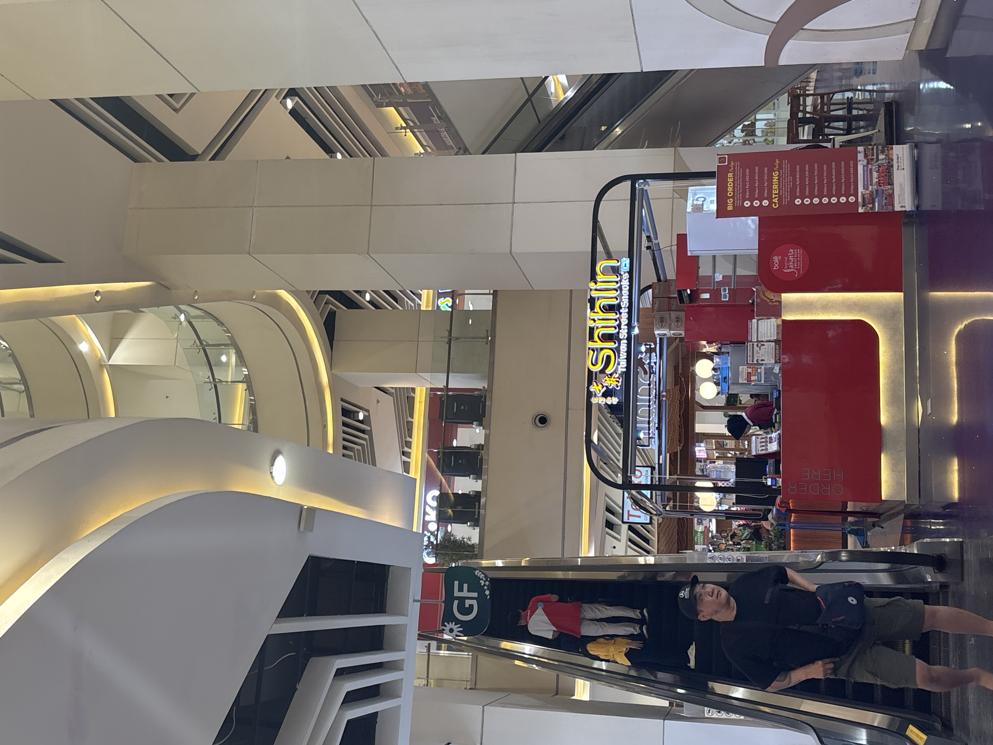 The image shows the interior of a mall in North Jakarta, Indonesia. In the foreground, a man in a black t-shirt, olive green shorts, and a black baseball cap is walking down an escalator, carrying a blue bag. To the right of the escalator is a red food kiosk with the text "Shihlin Taiwan Street Snacks" in yellow lettering. A sign on the kiosk lists prices for "BIG ORDER" and "CATERING" packages. In the background, another escalator is visible, as well as several levels of the mall's interior, including balconies and architectural features with ambient lighting. Several people can be seen on the escalators and within the mall.
