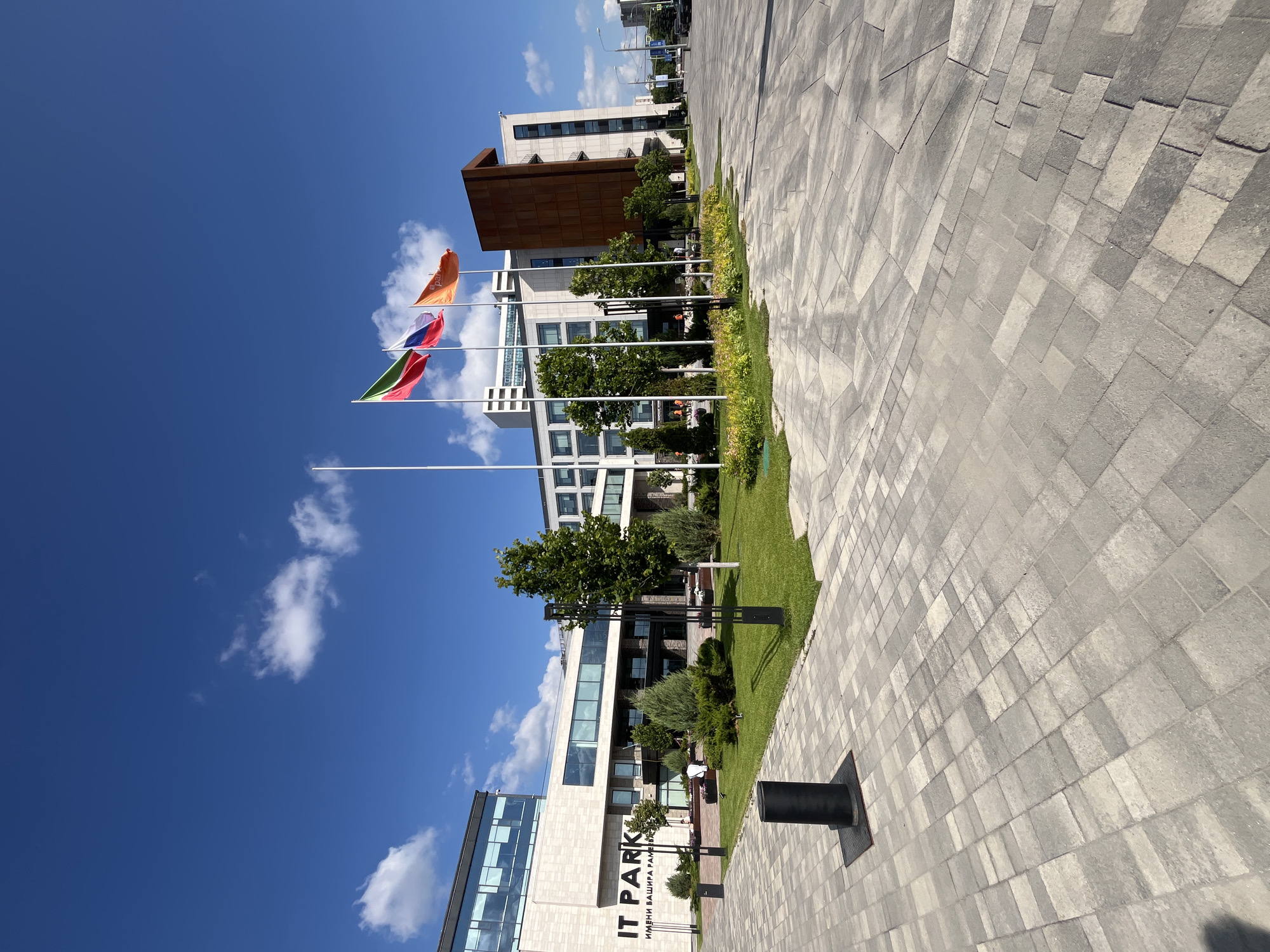 The image shows the IT Park building in Kazan, Russia, on a clear, sunny day. The building is a modern architectural complex with a light-colored facade and large windows. In front of the building, there is a paved plaza with a geometric pattern of grey and beige interlocking stones. A green lawn with scattered trees and flower beds is situated between the plaza and the building. Three flags are flying on flagpoles in front of the building: the flag of Tatarstan, the flag of Russia, and an orange flag with a logo. Several people are visible in the distance, walking or standing on the plaza and lawn. The overall scene depicts a well-maintained and modern urban environment.