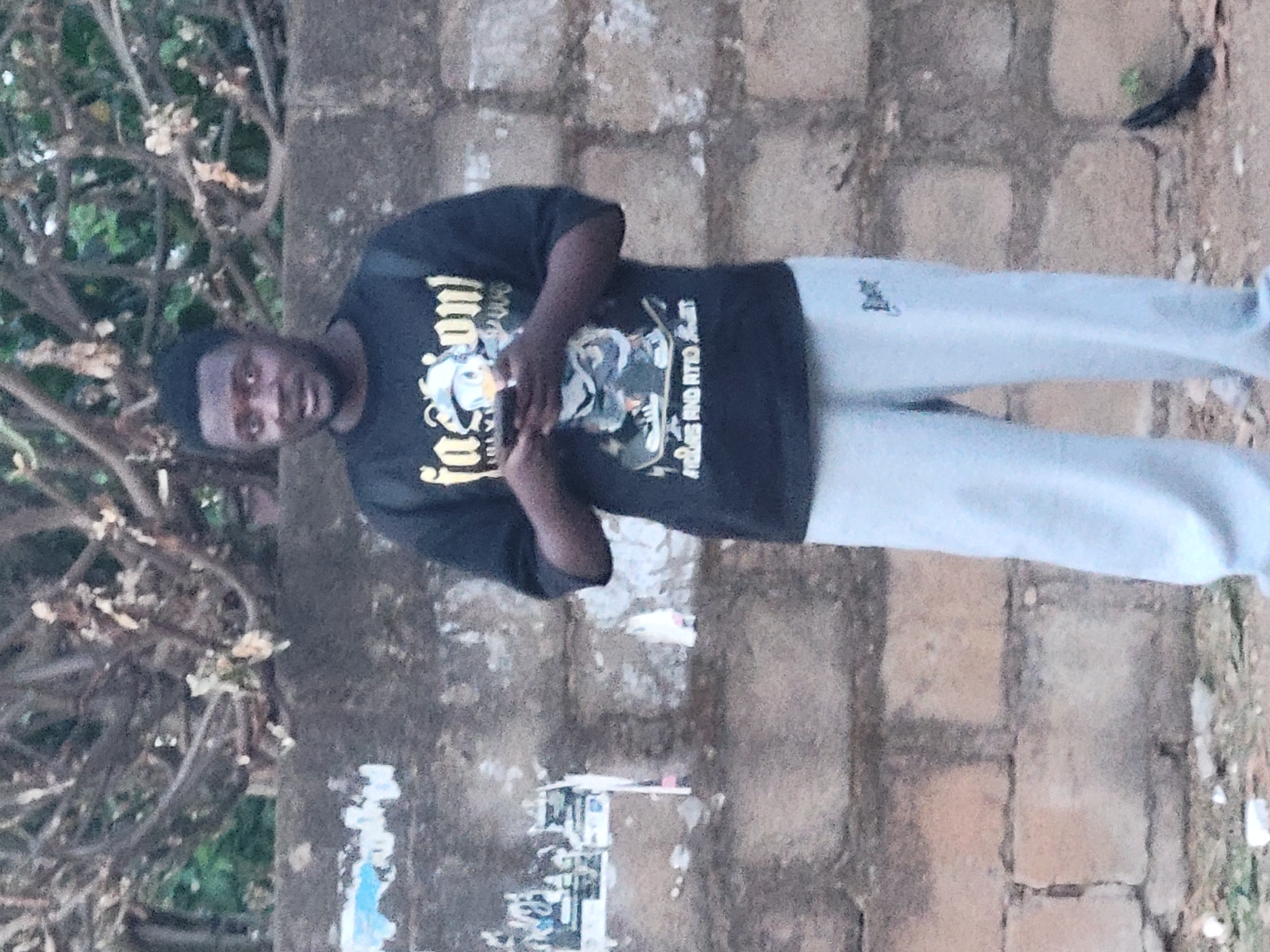 A man is standing outside in what appears to be Jos, Nigeria. The man is wearing a black t-shirt with a cartoon design, and grey sweatpants. He is standing in front of a concrete wall and some trees.
