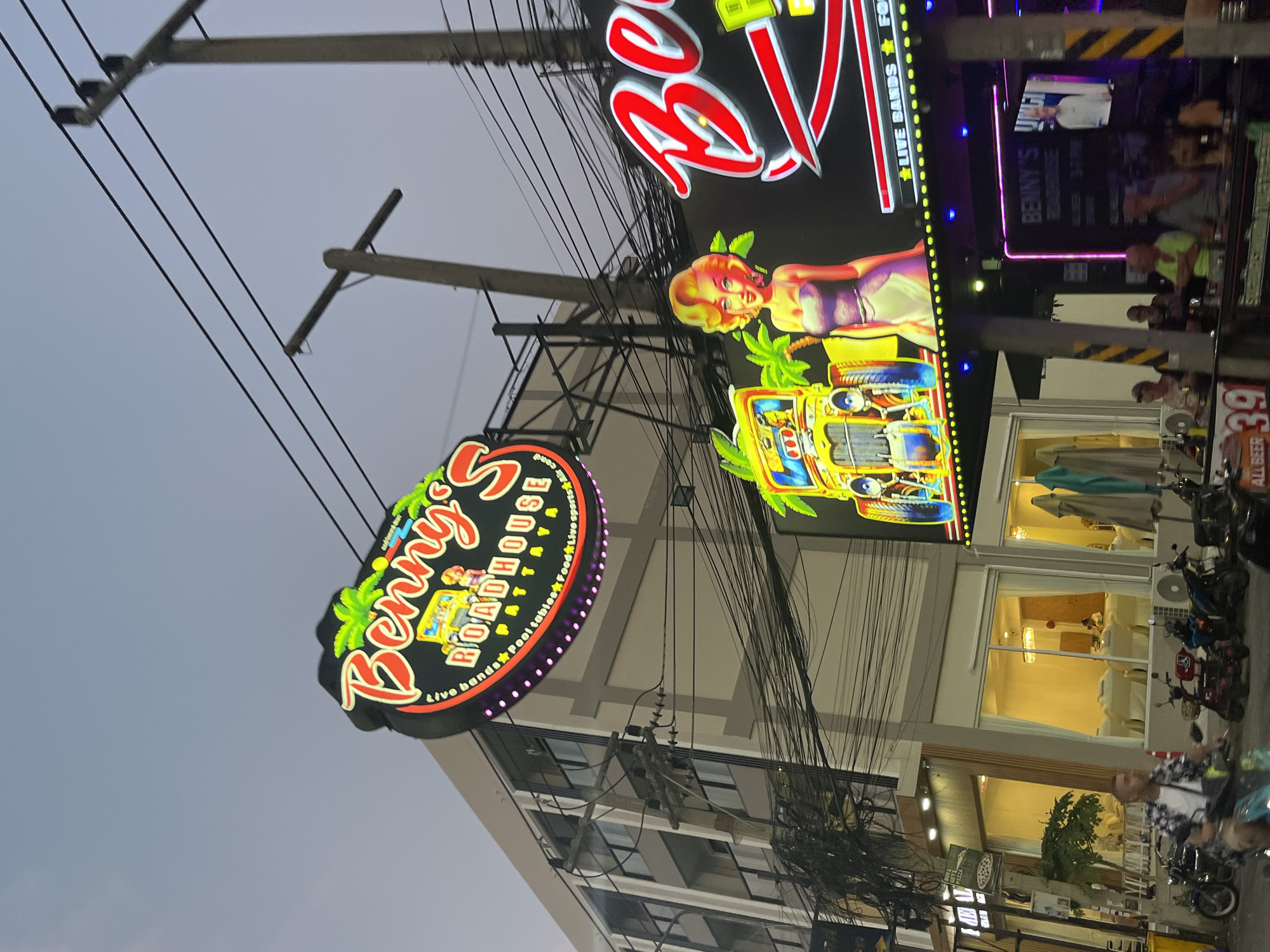 The image shows a street scene in Pattaya City, Thailand. Two brightly lit signs dominate the view: "Benny's Roadhouse Pattaya live sports Air cond, dol tables, foods," with a palm tree design and "Be... Benny's....live bands. The signs are suspended above a building, with visible electrical wires and a utility pole. The building has several lighted windows, revealing interior spaces. A group of people are visible on an outdoor balcony. Parked motorcycles line the street, with figures of people visible nearby.