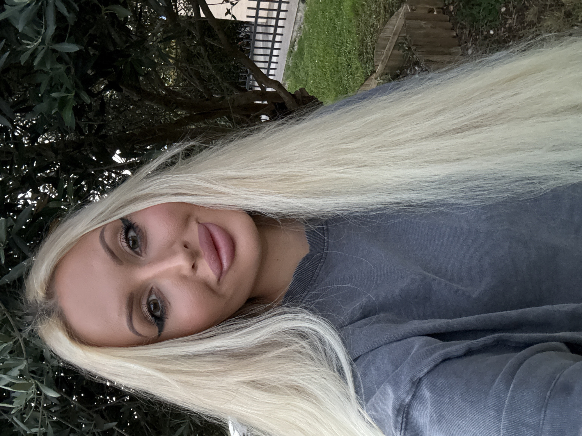 A selfie of a blonde woman with long, straight hair and heavy makeup is shown outdoors. She is wearing a grey t-shirt and is framed by greenery. The background shows a tree, a patch of grass, and a wooden fence. The overall lighting suggests it is daytime.