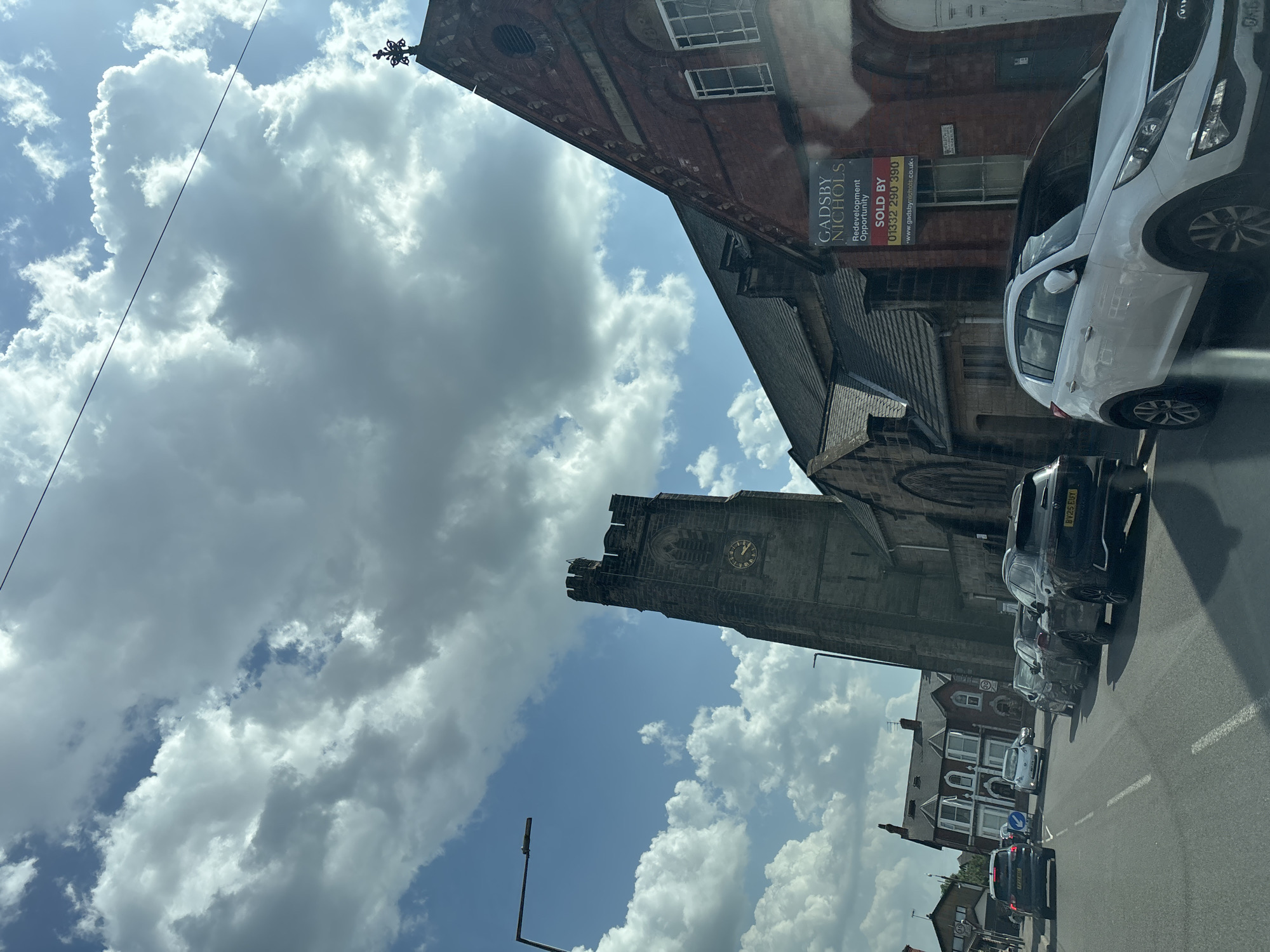 Captured from a vehicle's vantage point, the image depicts a street scene dominated by a historic church tower. The tower, constructed from gray stone, features a clock face and ornate architectural details. Adjacent to the church is a red brick building with signage indicating a "Redevelopment Opportunity," including a phone number. Cars are parked along the street, including white and black vehicles. The sky is filled with a mix of blue sky and large cumulus clouds. The presence of the church tower and building styles suggest the location is in Nottingham, United Kingdom.