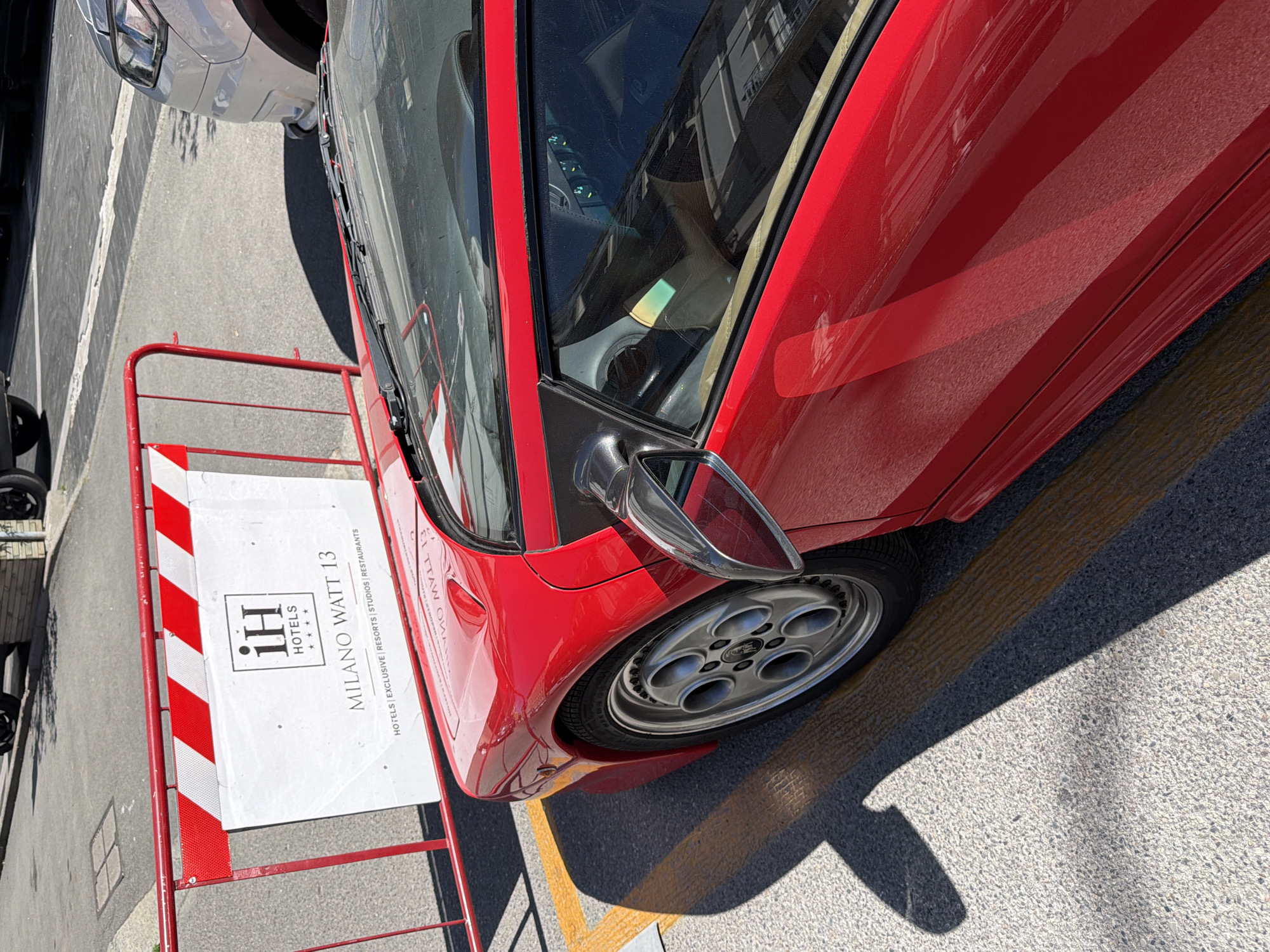 The image displays a vibrant red sports car, identifiable as a Lamborghini Diablo by its distinctive design and wheels, parked outdoors on a bright, sunny day. The car is positioned on an asphalt surface, marked with yellow lines, and a strong shadow is cast by the vehicle and other unseen objects, indicating clear skies and possibly midday.
To the immediate left of the car is a red and white striped barrier. Attached to this barrier is a prominent white sign. The sign features the "iH HOTELS" logo, along with four stars, and the text "MILANO WATT 13". Below this, it states "HOTELS | EXCLUSIVE | RESORTS | STUDIOS | RESTAURANTS". This clearly points to a location in Milan, Italy, specifically referencing the "Watt 13" area.
The red car's exterior is glossy, showing reflections of its surroundings. Its silver, multi-spoke wheels are visible, and through the tinted side window, reflections of buildings can be seen, along with a partial view of the car's dark interior and dashboard. A silver-grey car is partially visible in the background, parked further away. The scene captures a stationary vehicle in what appears to be a parking lot or street in an urban setting.