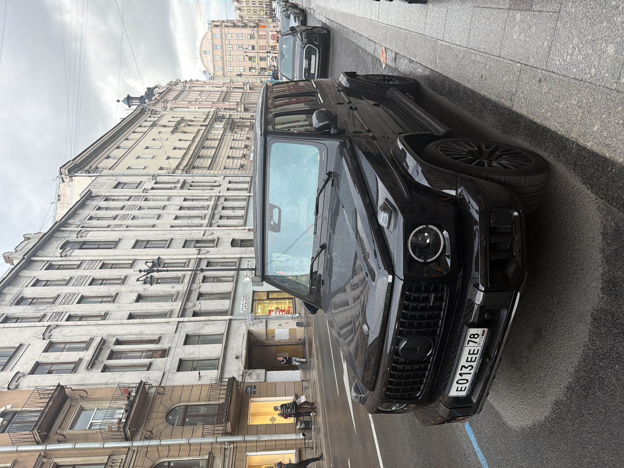 The image shows a black Mercedes-Benz G-Wagen parked on a wet city street. The car is angled towards the viewer, with its front grille and headlights prominently displayed. The license plate is visible and reads "E013EE 78 RUS".
In the background, there are tall, old European-style buildings with many windows. The architecture suggests an urban setting. There are also other cars parked further down the street.
A few people are visible in the distance, walking along the sidewalk and near shop entrances. One person is leaning against a wall. The ground appears wet, indicating that it has recently rained or is currently raining. The sky is overcast, suggesting a cloudy or dreary day. The overall mood is subdued, likely due to the weather and the dark color of the car. The scene appears to be a candid street photograph, capturing a moment in an urban environment.