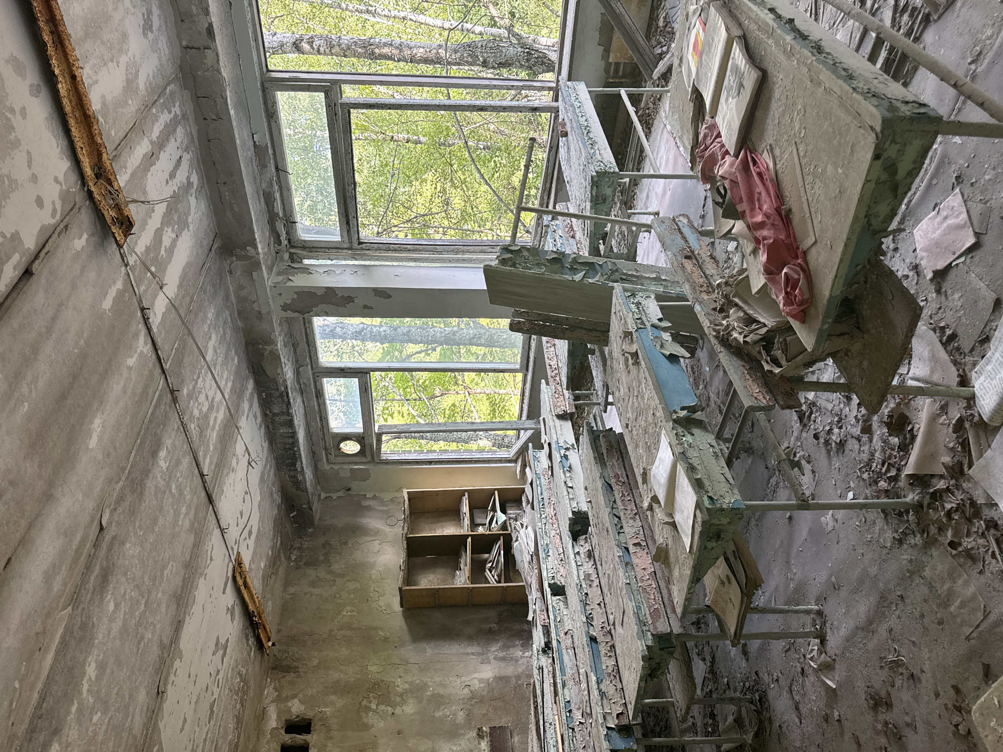 This media file captures the interior of a derelict classroom or study hall. The scene is characterized by decay and abandonment. The walls are stained and peeling, revealing concrete beneath layers of paint. Large windows, some broken or cracked, allow natural light to filter in, illuminating the dust-laden air. Outside the windows, lush green trees suggest an overgrown, possibly temperate environment, and hint at the time of year being spring or summer.
The room is furnished with rows of old desks or tables, their surfaces covered in dust and debris. Some of these surfaces are still occupied by books, their pages weathered and possibly water-damaged. A crumpled piece of pink fabric, perhaps clothing, lies on one of the desks, adding a touch of human residue to the desolate scene. A simple wooden bookshelf with multiple compartments stands against one wall, containing more scattered books and papers. The floor is littered with fallen leaves, dirt, and fragments of paper, indicating a significant passage of time since the room was last used.
The overall atmosphere is one of profound neglect and stillness. There are no people visible, and the objects left behind suggest a hasty departure or a slow surrender to the elements. The play of light and shadow, the textures of decay, and the presence of nature encroaching on the man-made structure create a melancholic and haunting visual narrative. The time of day appears to be daytime, with the natural light suggesting it could be morning or afternoon. There are no overt clues to the specific location other than the environmental setting visible through the windows. No visible text is discernable.