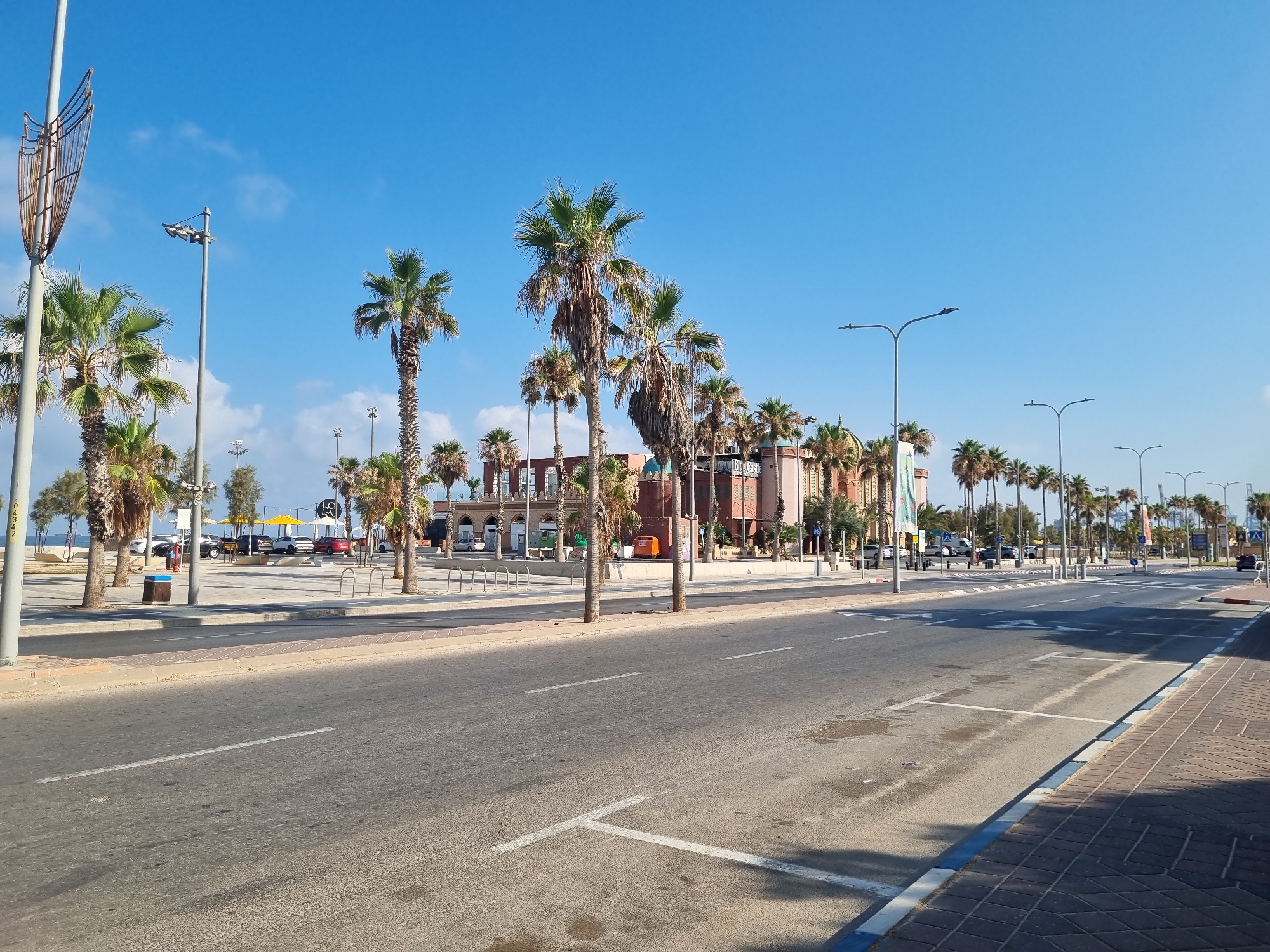 The image captures a street scene in Ashdod, Israel, likely during daylight under a clear blue sky. The foreground features a two-lane asphalt road with white markings indicating lanes and parking spaces. To the right, a brick-patterned sidewalk runs alongside the road, bordered by a blue and white curb. Beyond the sidewalk, a line of palm trees creates a natural barrier. In the mid-ground, a promenade area is visible with parked cars, some under yellow awnings. Buildings with red-brick facades and arched windows, possibly restaurants or shops, line this area, with palm trees interspersed. Several streetlights and traffic lights are visible along the road and promenade. The overall environment suggests a coastal city, with the presence of palm trees and the bright, open sky reinforcing this impression. There are no visible people actively engaged in any interactions or actions.